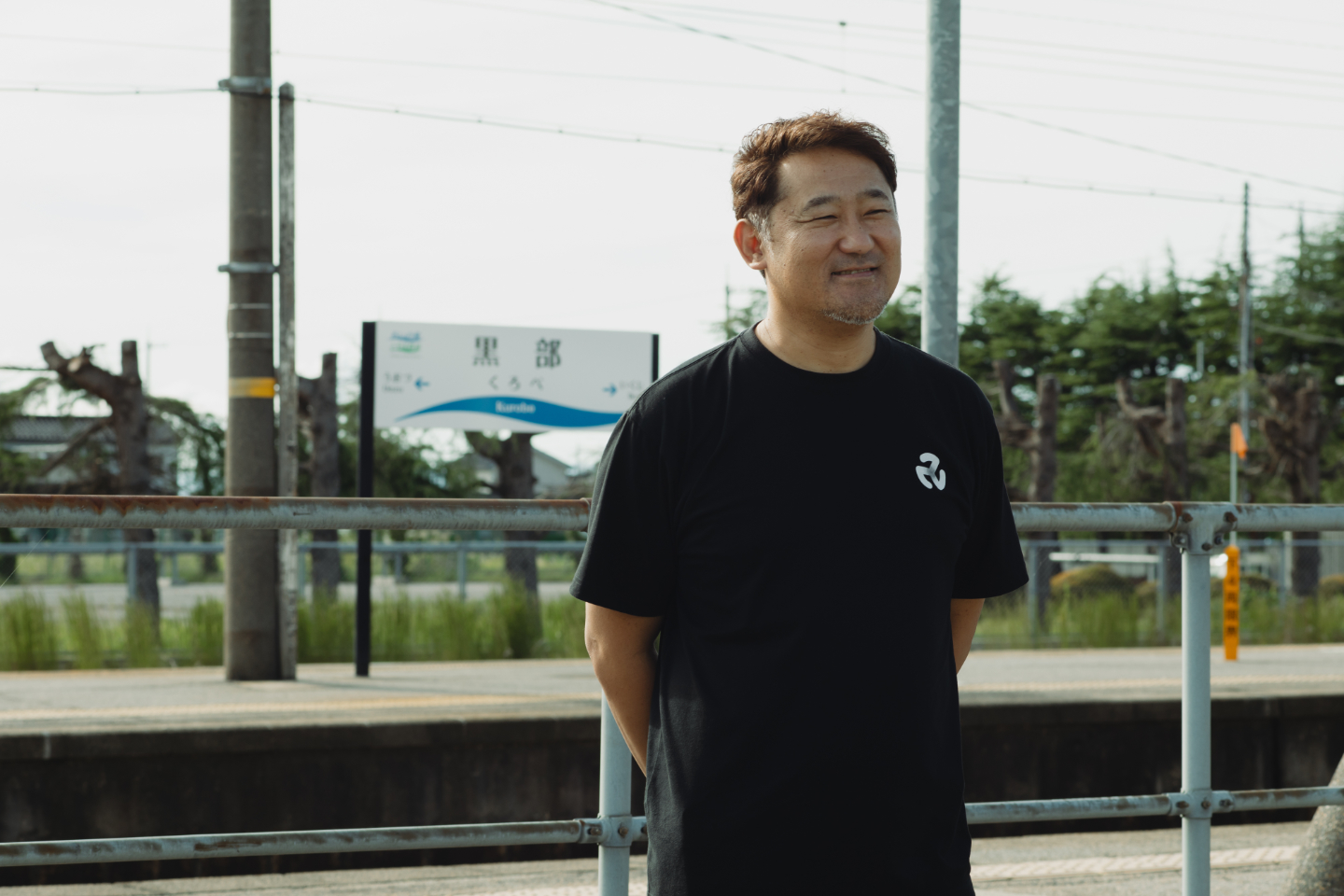【写真】駅のホームで小柴さんが少し横を向いて微笑んで立っている。黒部と書かれた駅名標が背景に見える