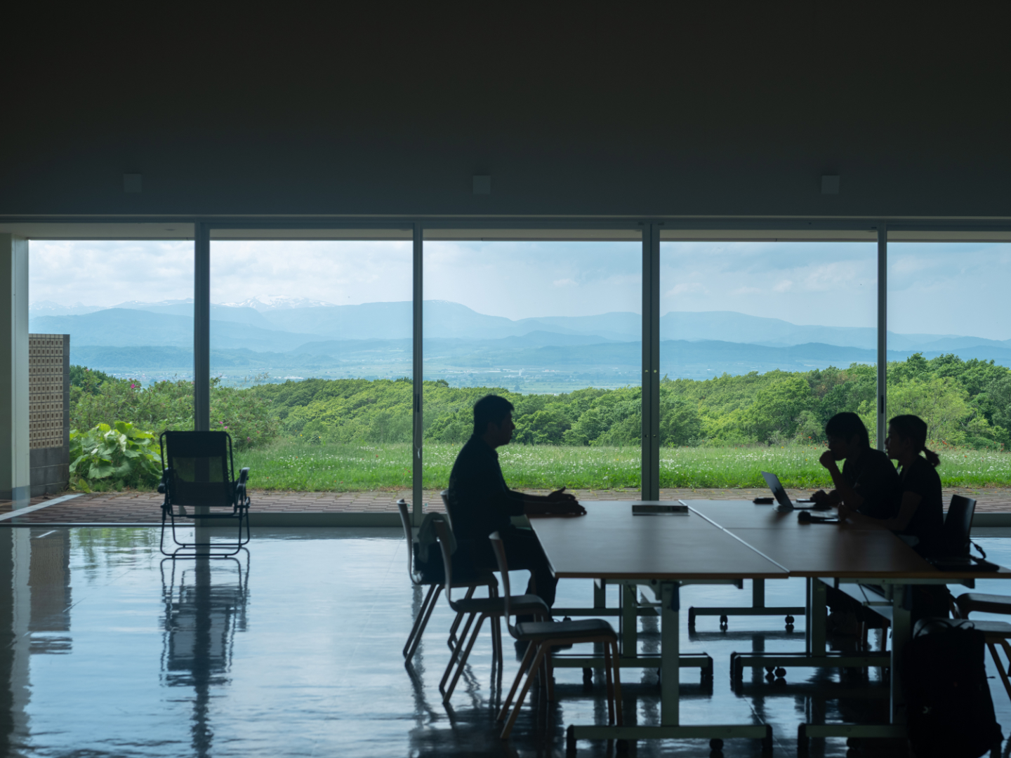 【写真】大きな窓のある建物の中で、広いテーブルの片側に1人、もう片側に2人が座り向き合って話し込む。背景には緑と空の大自然