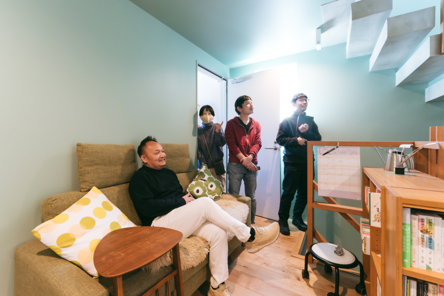 【写真】緑色の壁とのある空間で、一人がソファに座り、他の3人が本棚や階段を眺めている