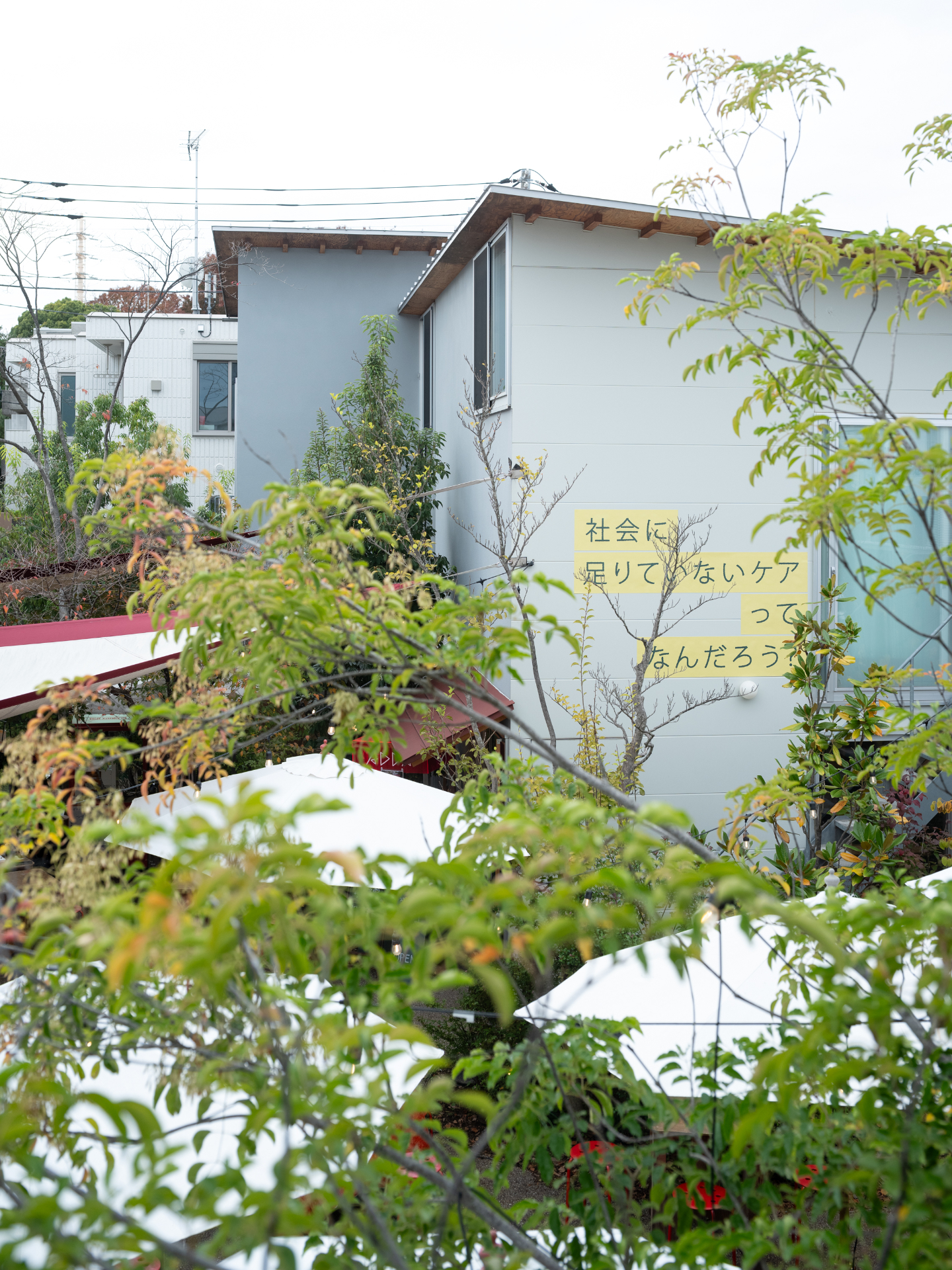 【写真】木や植物に囲まれた建物の外観、外壁に日本語のメッセージが描かれています。壁には「社会に足りていないケアってなんだろう」の言葉