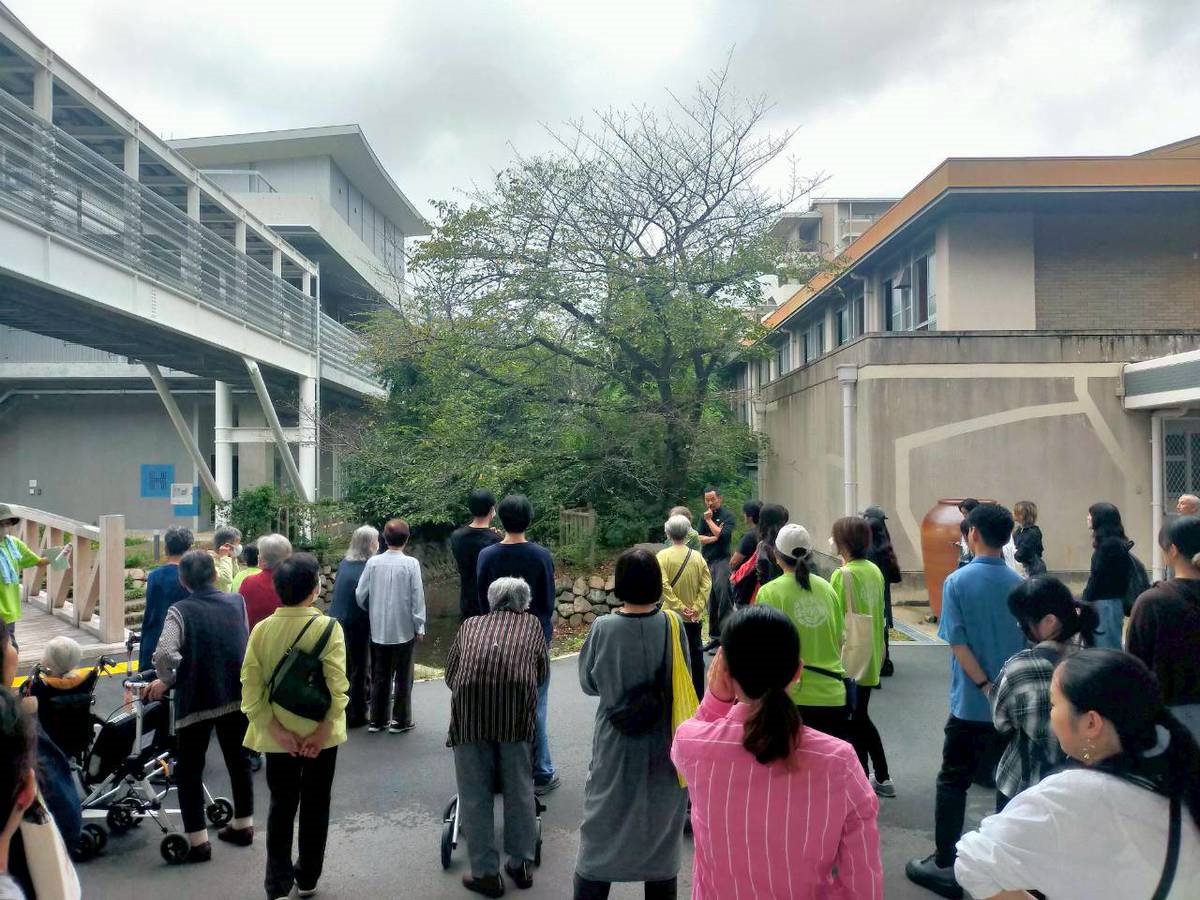 【画像】建物と建物の間にある大きな木を囲んで十数名の人が話を聞いている