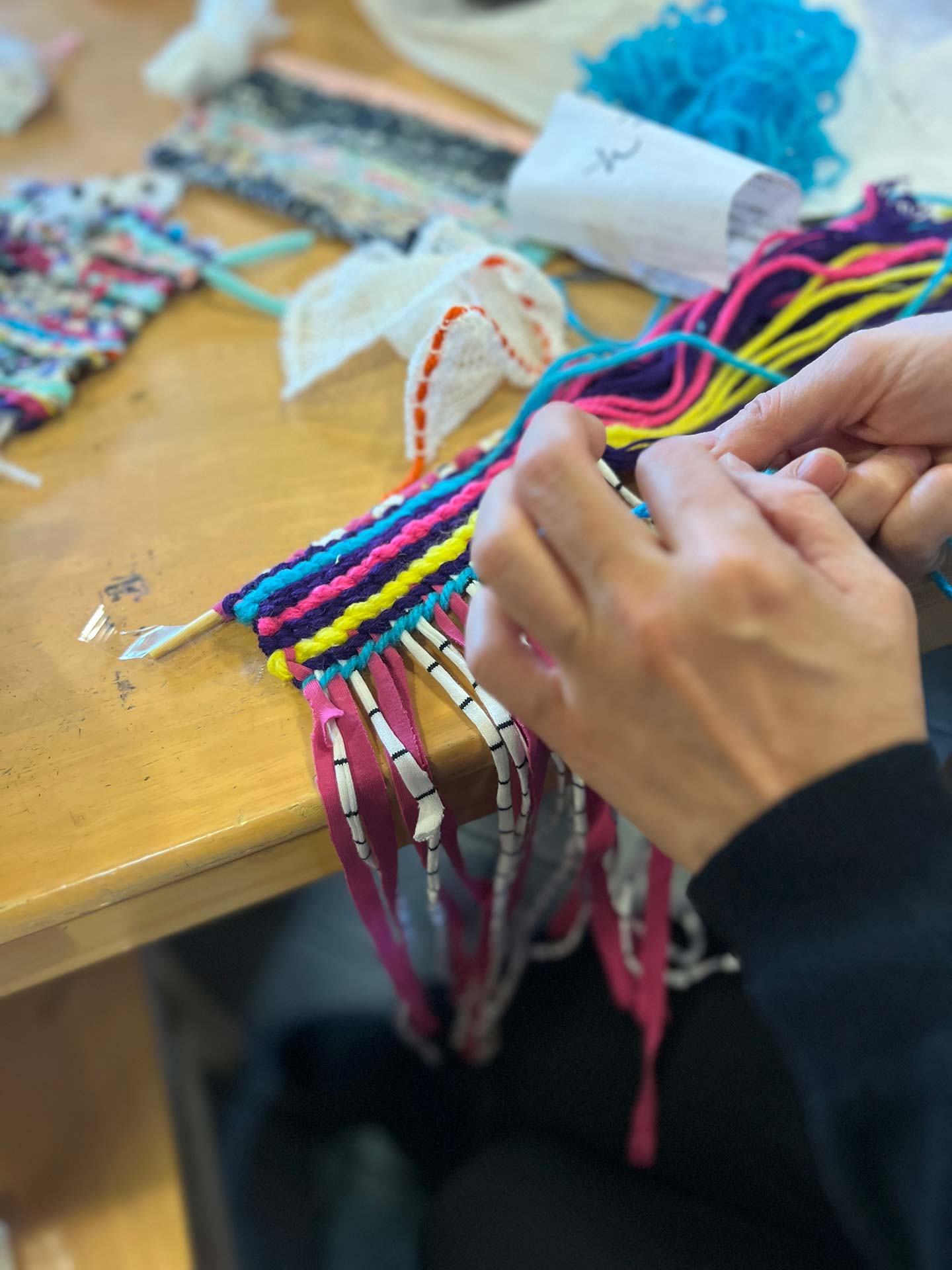 ピンク、黄色、青、黒などの横糸でカラフルな布を織る人の手元写真。