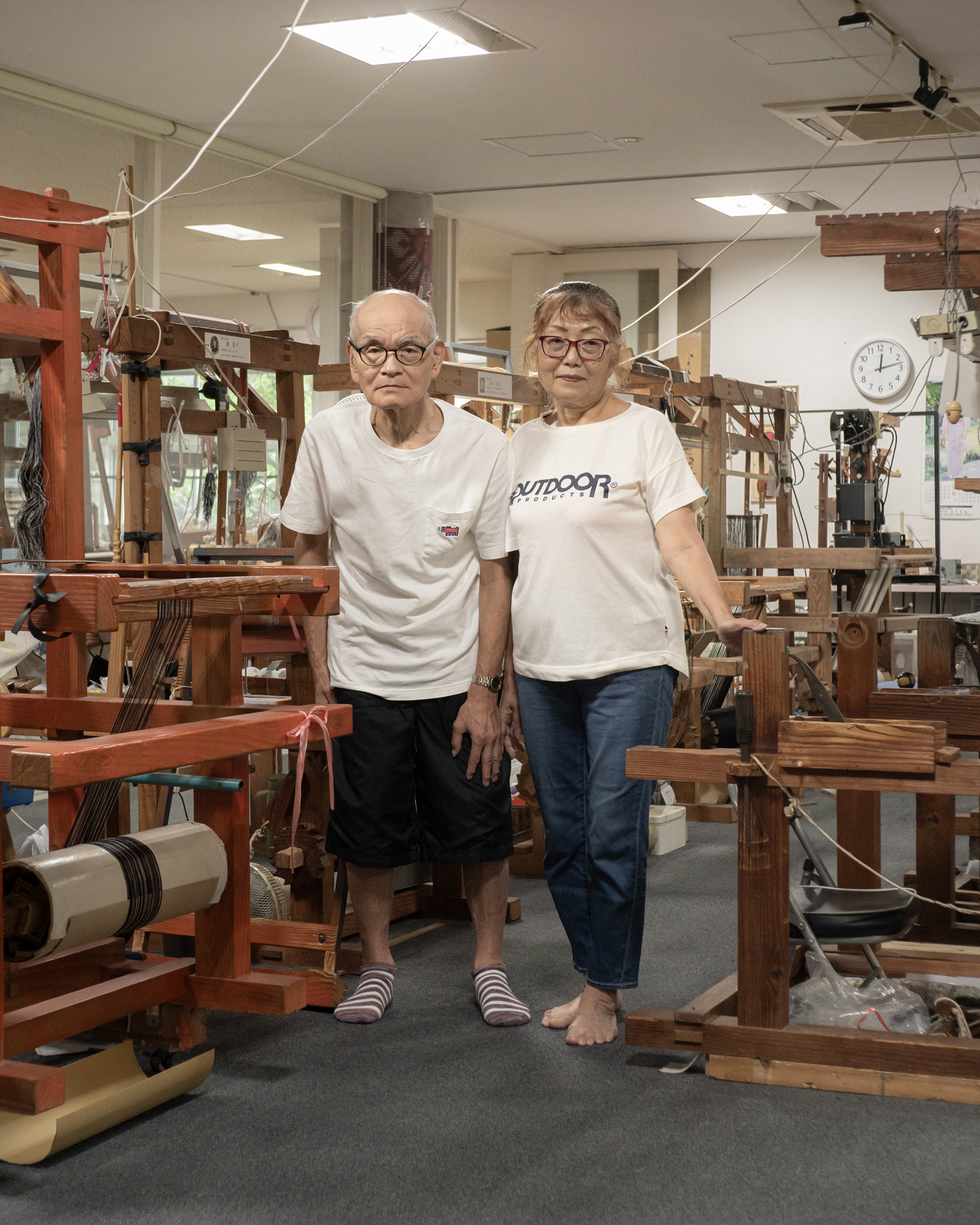 【写真】室内に木製の機織り機がたくさん並んでいる。画面中央に池田さん夫妻が立っている。左側が夫、右側が妻。ふたりとも白い半袖のTシャツを着ていてメガネをかけている。夫は黒いハーフパンツにしましまの靴下、妻はデニムパンツで素足。
