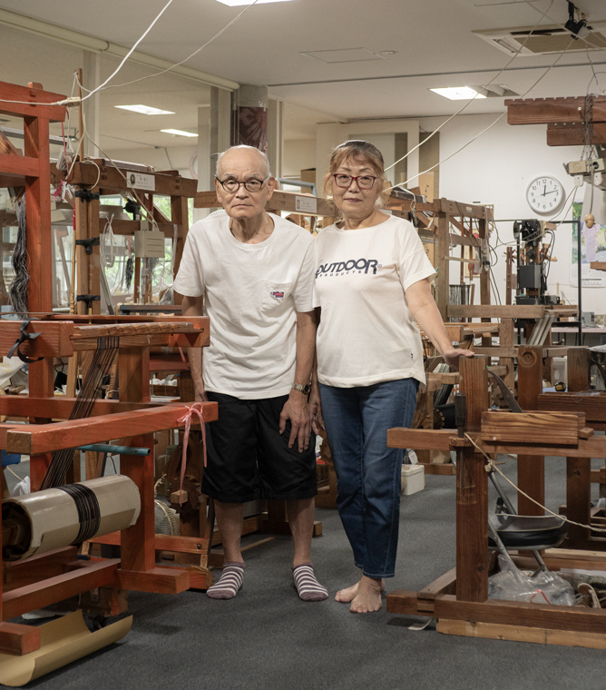 【写真】室内に木製の機織り機がたくさん並んでいる。画面中央に池田さん夫妻が立っている。左側が夫、右側が妻。ふたりとも白い半袖のTシャツを着ていてメガネをかけている。夫は黒いハーフパンツにしましまの靴下、妻はデニムパンツで素足。