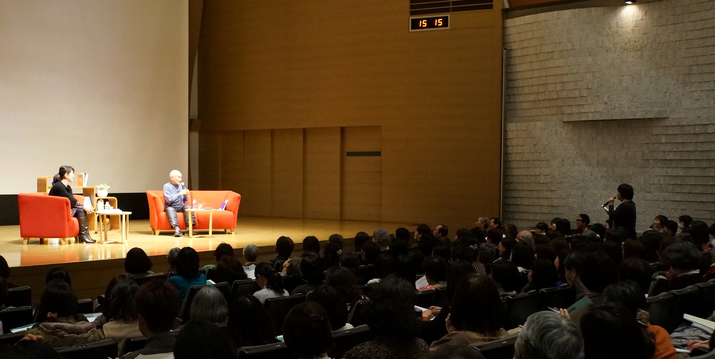 【写真】広いホール。ステージの上で2人がオレンジのソファに座って話している