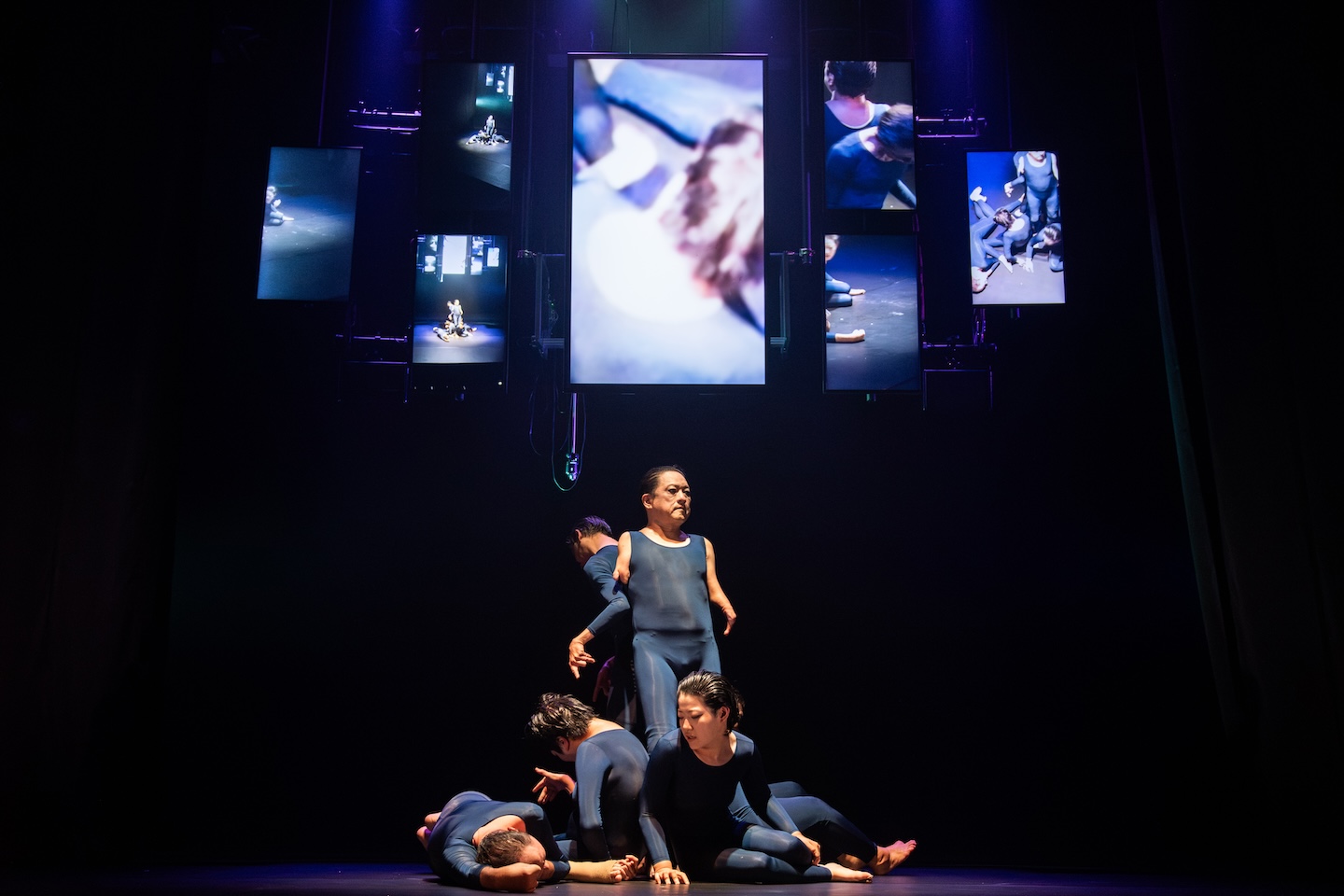 映像が映し出された6つのパネルの前に、青いレオタードを身に着けた5人の人が集まってポーズをとっている