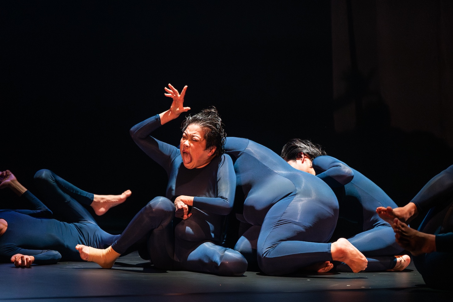 青いレオタードを身に着けた人が寝転んだり座ったりしている。真ん中の人は右手を頭の上にあげ、大きく口を開いている