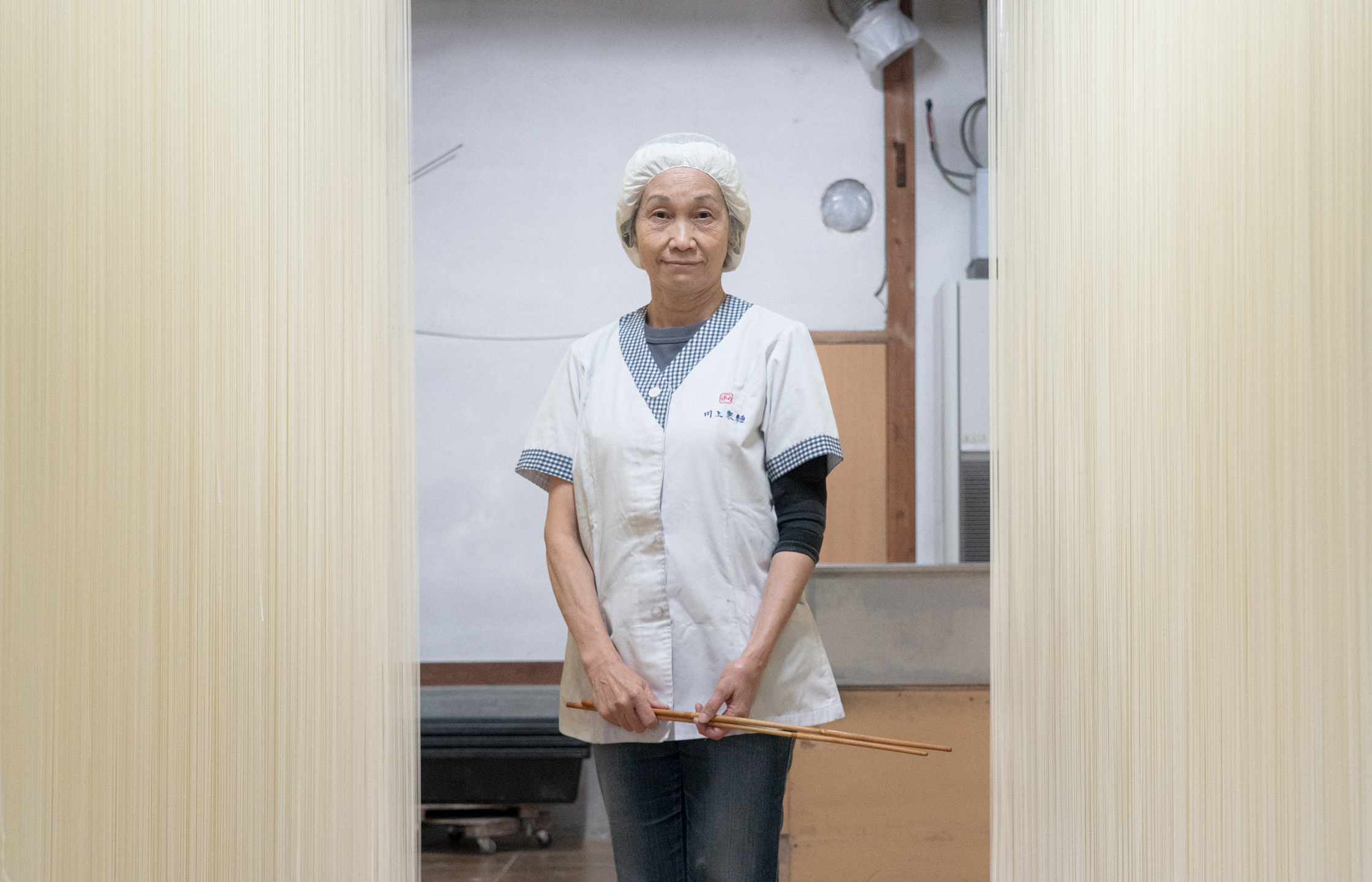 【写真】川上製麺の制服を着たいざきじゅんこさんが中央に立っている縦長の写真。半袖の白い前ボタンの制服に、白い帽子をかぶっている。デニムパンツと白いスニーカーも履いている。両手を前に揃えていて手には長い箸を一膳もっている。いざきさんの両側を挟むように、天井から床までびっしりと手延べそうめんが干されている。