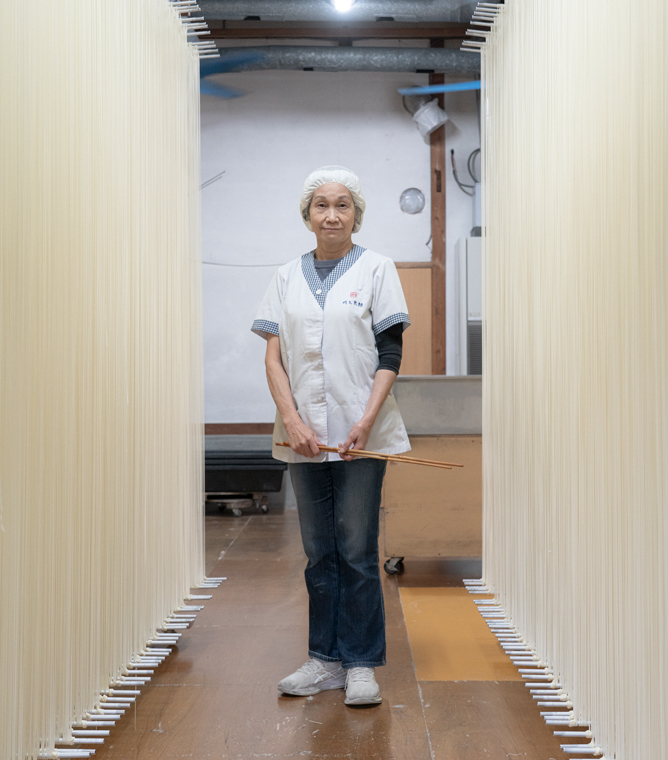 【写真】川上製麺の制服を着たいざきじゅんこさんが中央に立っている縦長の写真。半袖の白い前ボタンの制服に、白い帽子をかぶっている。デニムパンツと白いスニーカーも履いている。両手を前に揃えていて手には長い箸を一膳もっている。いざきさんの両側を挟むように、天井から床までびっしりと手延べそうめんが干されている。