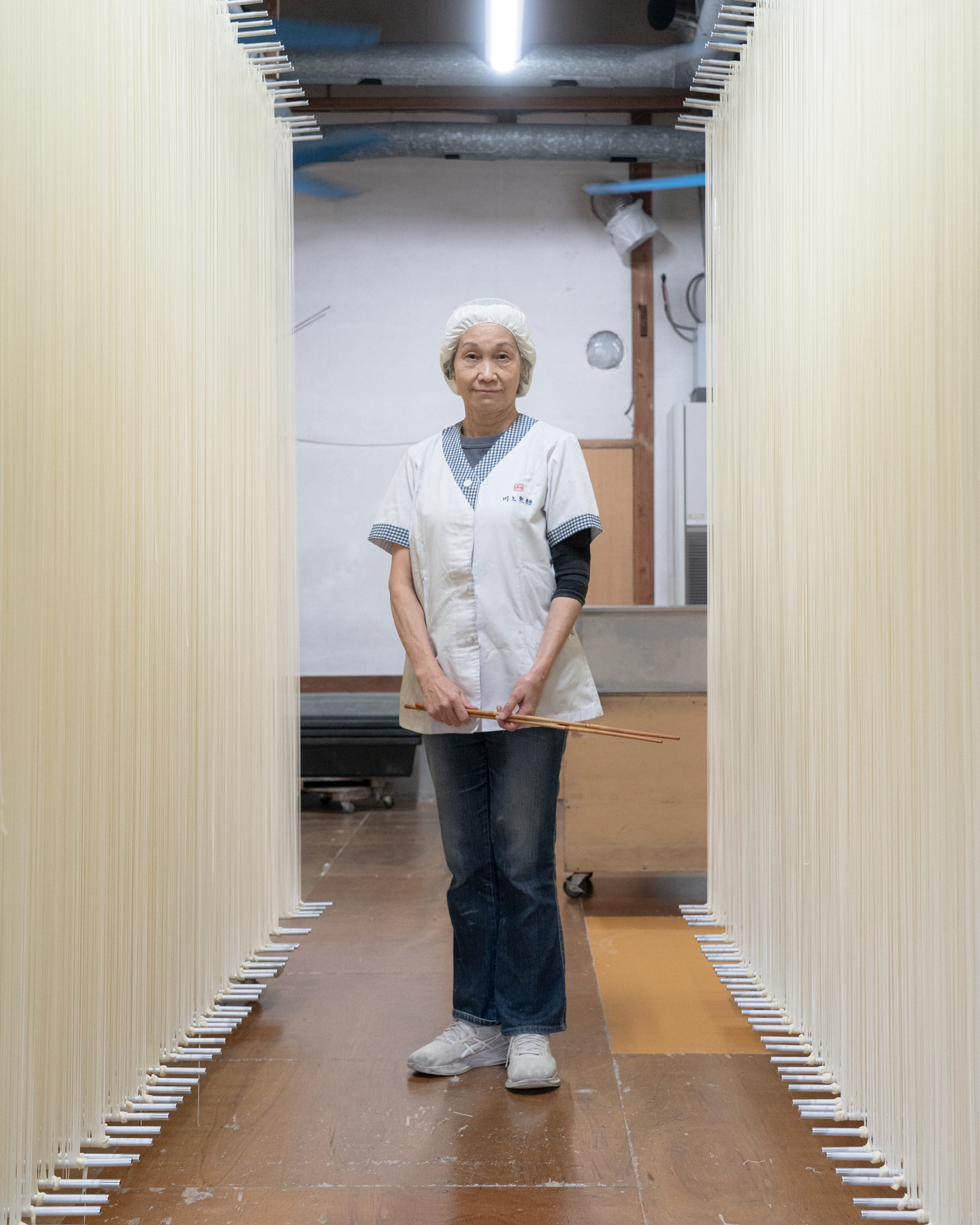 【写真】川上製麺の制服を着たいざきじゅんこさんが中央に立っている縦長の写真。半袖の白い前ボタンの制服に、白い帽子をかぶっている。デニムパンツと白いスニーカーも履いている。両手を前に揃えていて手には長い箸を一膳もっている。いざきさんの両側を挟むように、天井から床までびっしりと手延べそうめんが干されている。