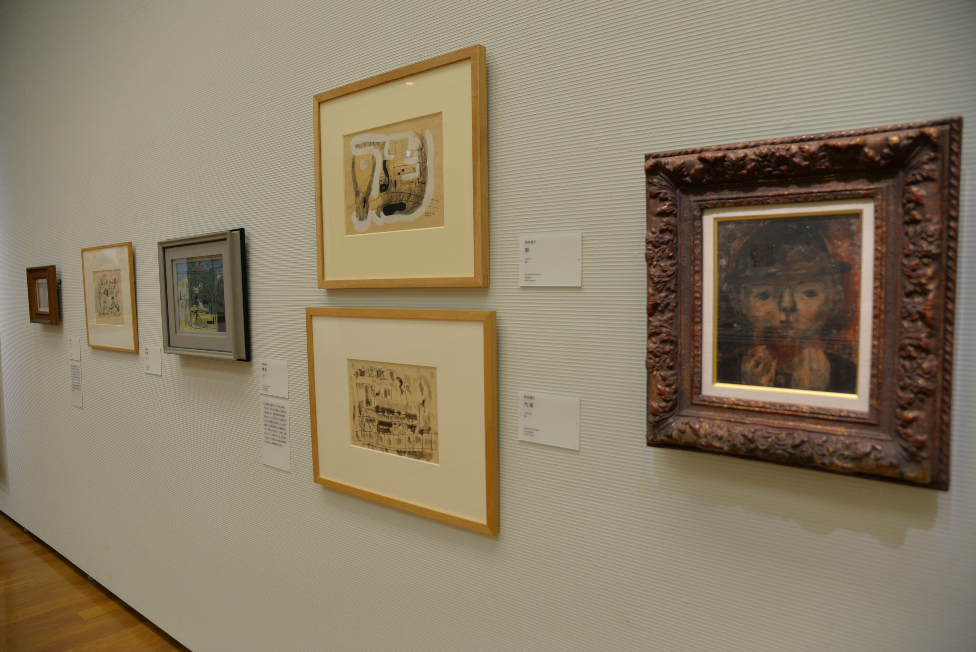 【写真】壁にずらりと額装された絵画が並ぶ。手前に、展覧会のチラシトップの絵