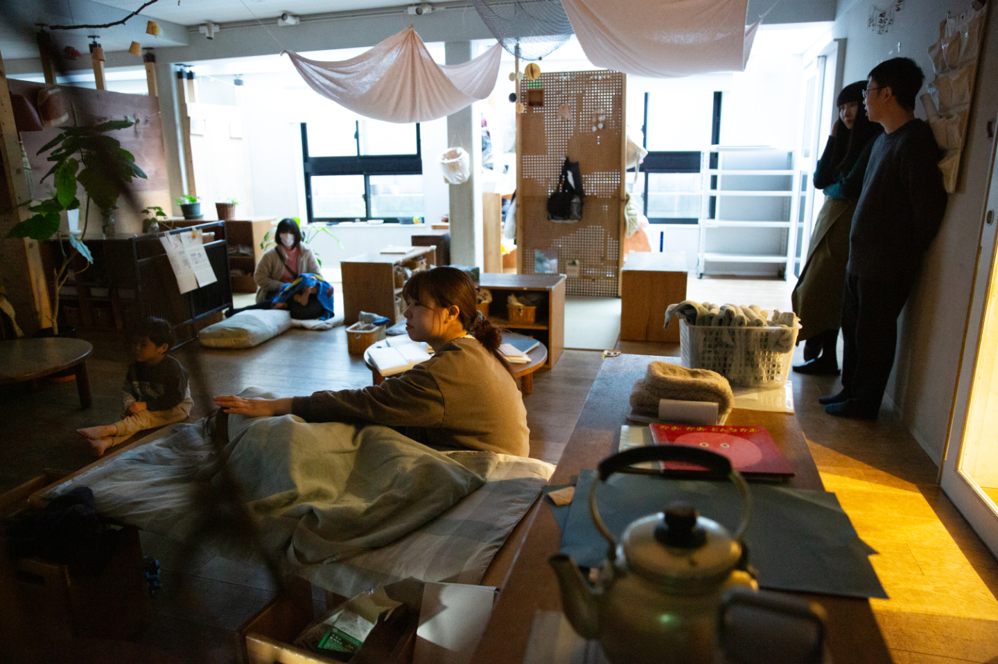 【写真】天井から布が吊るされ、木製家具や植物が配置された温かみのある空間。やわらかな照明に包まれた室内で、大人と子どもが床に座ってくつろぎ、横で青山さん寺尾さんが立ち話をしていいる