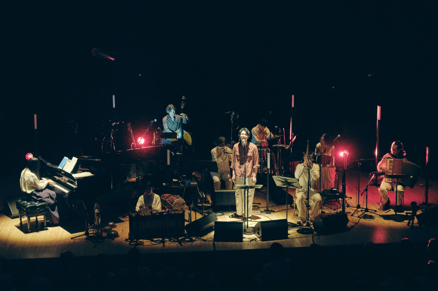 【写真】暗いステージ上で、赤い照明に照らされながらバンドが演奏している。中央にはボーカルが立ち、ピアノやドラム、ベース、アコーディオンなどの演奏者が並ぶ