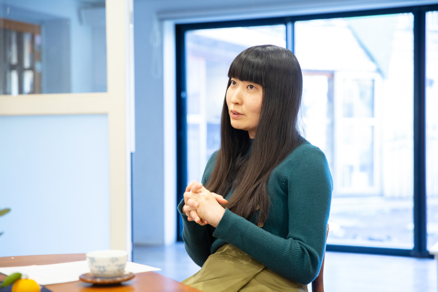 【写真】明るい室内で、テーブルに座り両手を軽く組みながら話す寺尾さん。窓から自然光が差し込み、テーブルにはコップや資料が置かれている