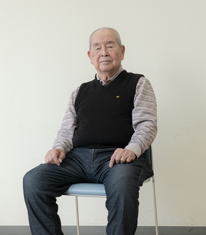 【写真】白い壁の室内で椅子に座って正面を見ている岡本ゆうすけさん。黒いニットベストの下、ボーダーの襟付きシャツをきて、デニムパンツに白いスニーカーを履いている。髪は白髪で膝の上に手を載せている。