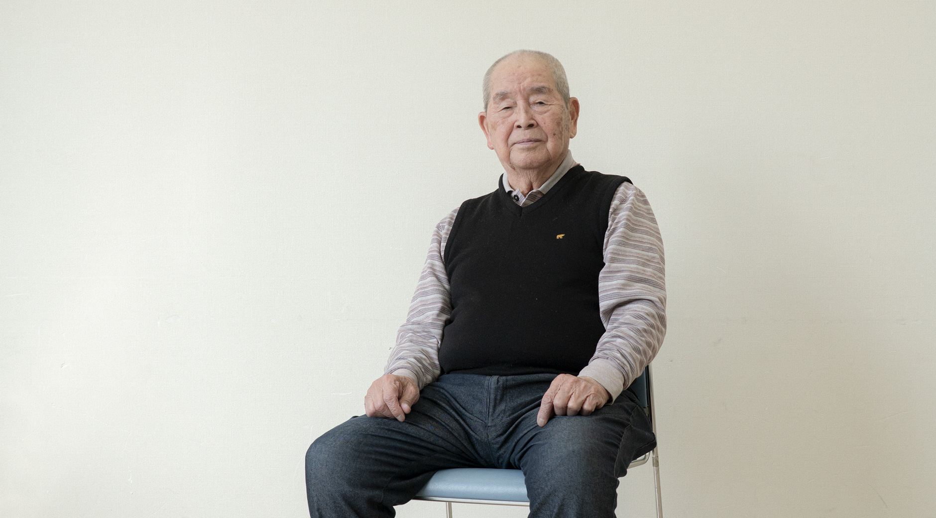 【写真】白い壁の室内で椅子に座って正面を見ている岡本ゆうすけさん。黒いニットベストの下、ボーダーの襟付きシャツをきて、デニムパンツに白いスニーカーを履いている。髪は白髪で膝の上に手を載せている。