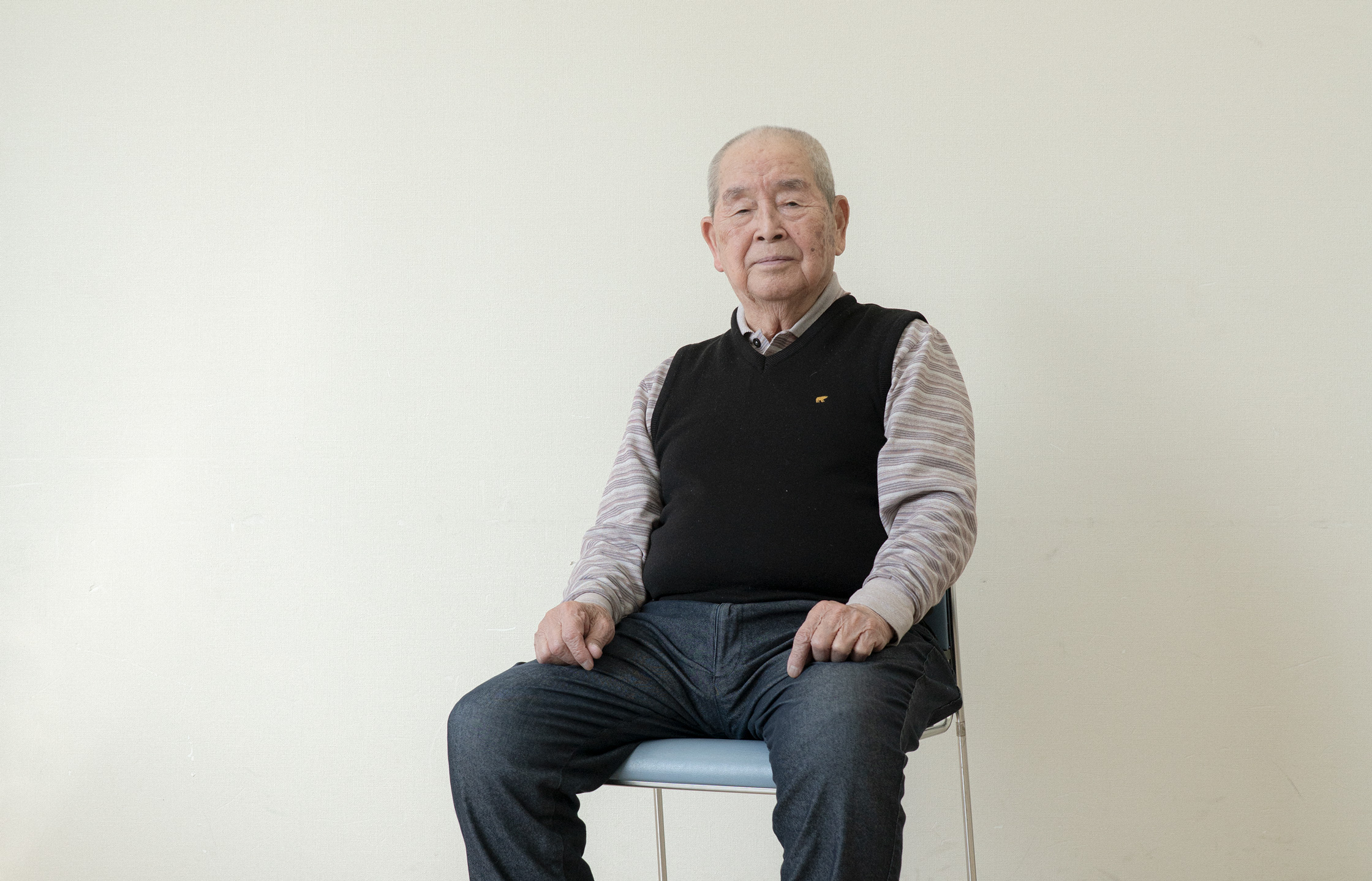 【写真】白い壁の室内で椅子に座って正面を見ている岡本ゆうすけさん。黒いニットベストの下、ボーダーの襟付きシャツをきて、デニムパンツに白いスニーカーを履いている。髪は白髪で膝の上に手を載せている。