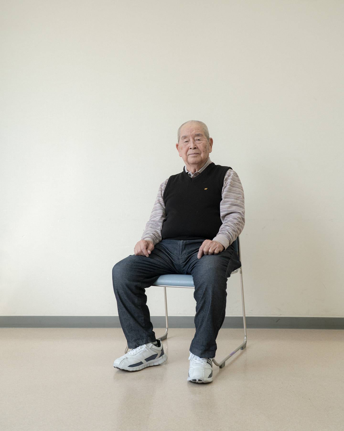 【写真】白い壁の室内で椅子に座って正面を見ている岡本ゆうすけさん。黒いニットベストの下、ボーダーの襟付きシャツをきて、デニムパンツに白いスニーカーを履いている。髪は白髪で膝の上に手を載せている。