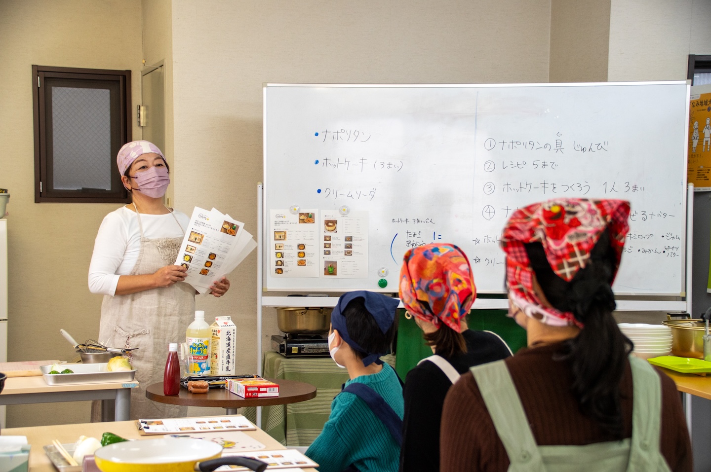 ホワイトボードの横にレシピを手にして立っている人、その人の話を座って聞いてる様子の人たちが手前に3人いる