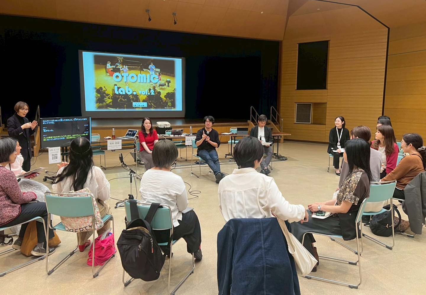 「OTOMIC Lab. vol.2」のラウンドテーブルディスカッションの様子。ホールのステージにあるスクリーンには「OTOMIC Lab. vol.2」と書いてある。年齢も性別もさまざまな人が円になって椅子に座っている。