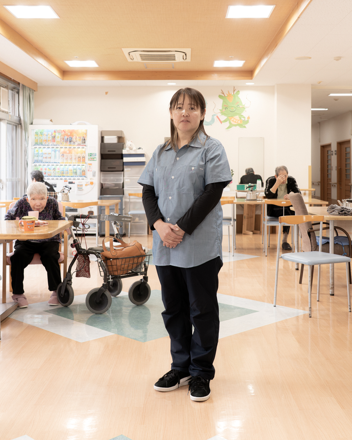 【写真】施設の中で立つおおばやしみかさんの写真。半袖のデニムシャツの下に長い黒色のTシャツ、黒いパンツをきている。髪の毛は肩までかっかっていて手を前で揃えて正面を見てたっている。背景には、ホームを利用する高齢者の人たちが座っている。