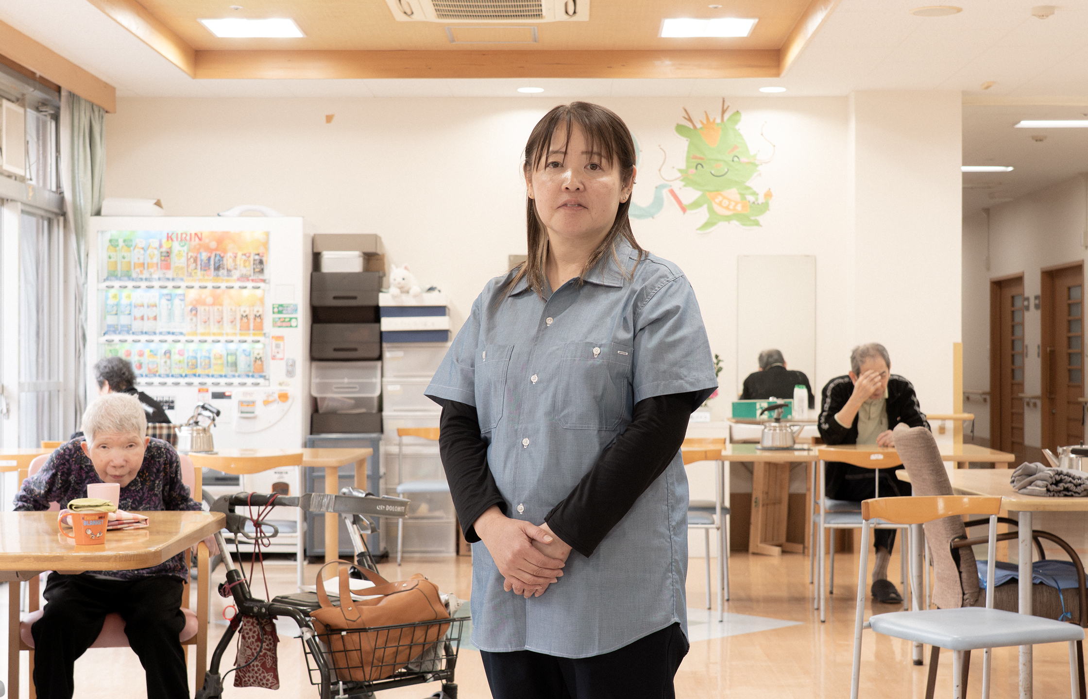 【写真】施設の中で立つおおばやしみかさんの写真。半袖のデニムシャツの下に長い黒色のTシャツ、黒いパンツをきている。髪の毛は肩までかっかっていて手を前で揃えて正面を見てたっている。背景には、ホームを利用する高齢者の人たちが座っている。