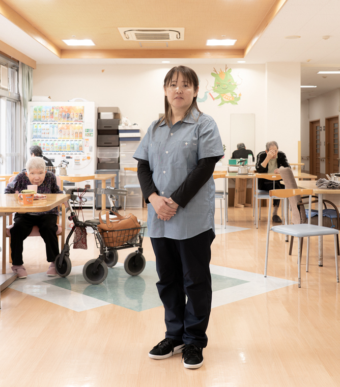 【写真】施設の中で立つおおばやしみかさんの写真。半袖のデニムシャツの下に長い黒色のTシャツ、黒いパンツをきている。髪の毛は肩までかっかっていて手を前で揃えて正面を見てたっている。背景には、ホームを利用する高齢者の人たちが座っている。