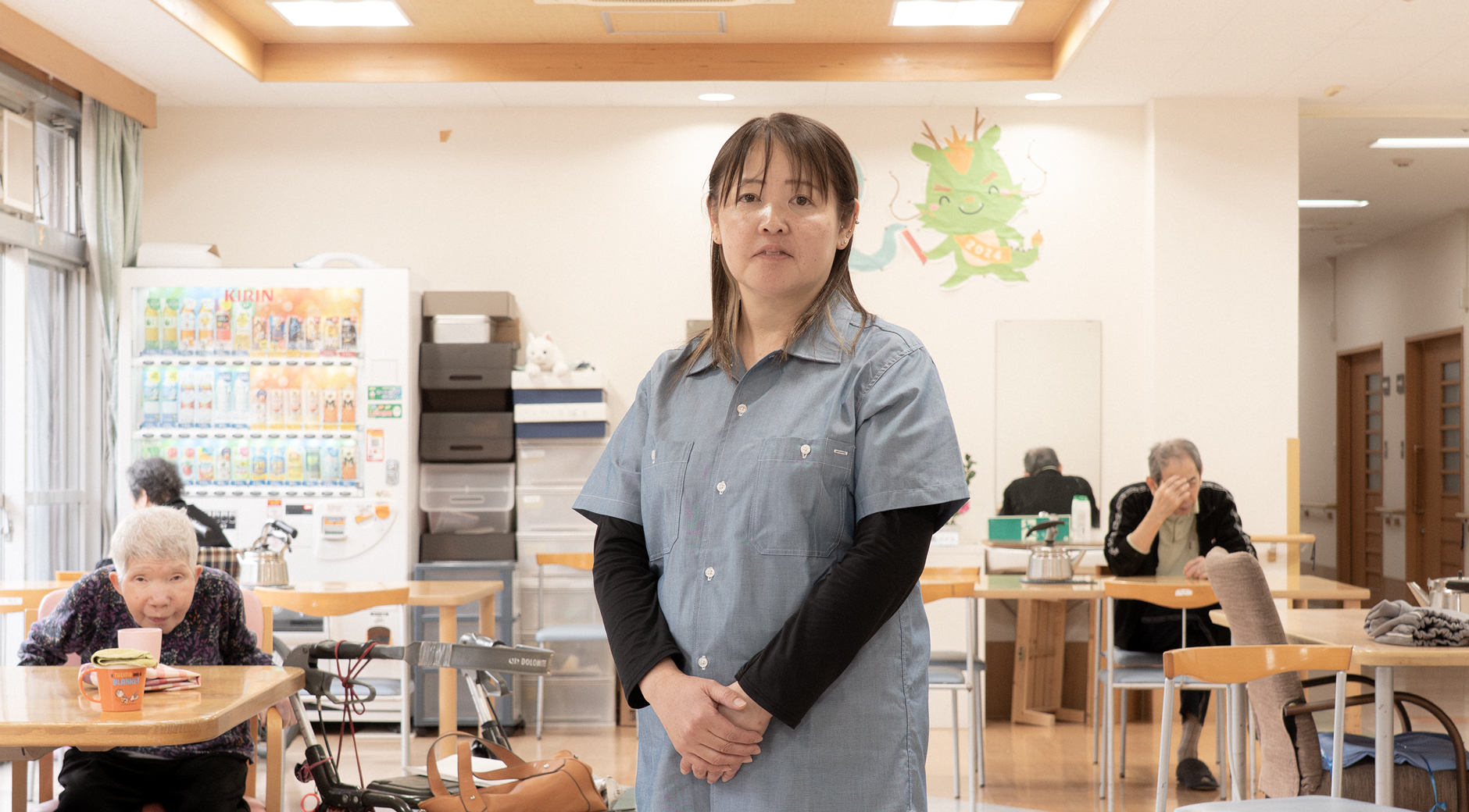 【写真】施設の中で立つおおばやしみかさんの写真。半袖のデニムシャツの下に長い黒色のTシャツ、黒いパンツをきている。髪の毛は肩までかっかっていて手を前で揃えて正面を見てたっている。背景には、ホームを利用する高齢者の人たちが座っている。