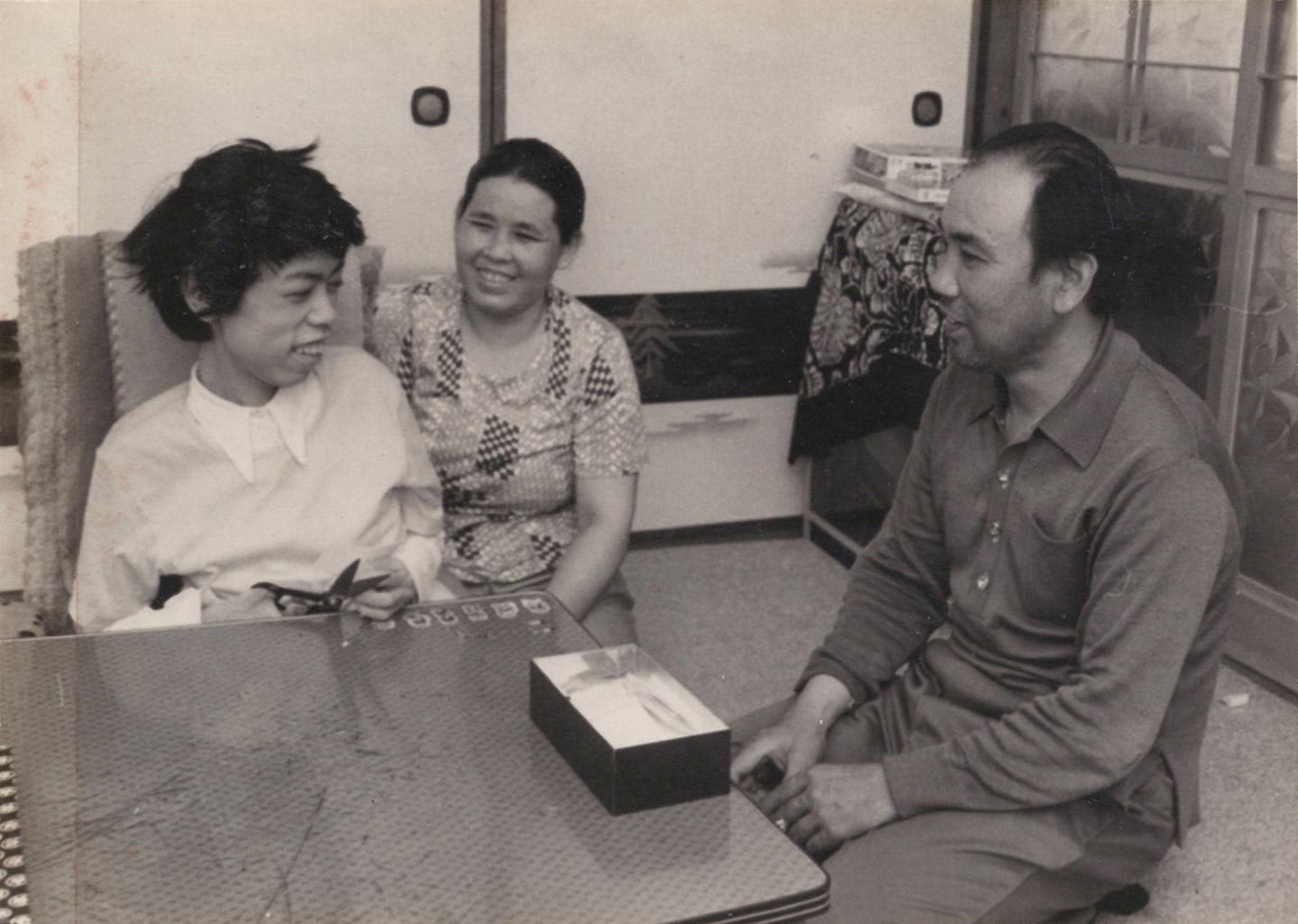 写真。座卓を前に座る聡さん。手には裁ちばさみが。横には父の平安喜さん、後ろには母が寄り添い、笑顔で写っている