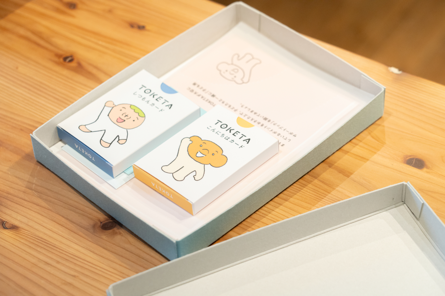 【写真】箱に収められた、カードセットと冊子が木製テーブルの上に置かれている