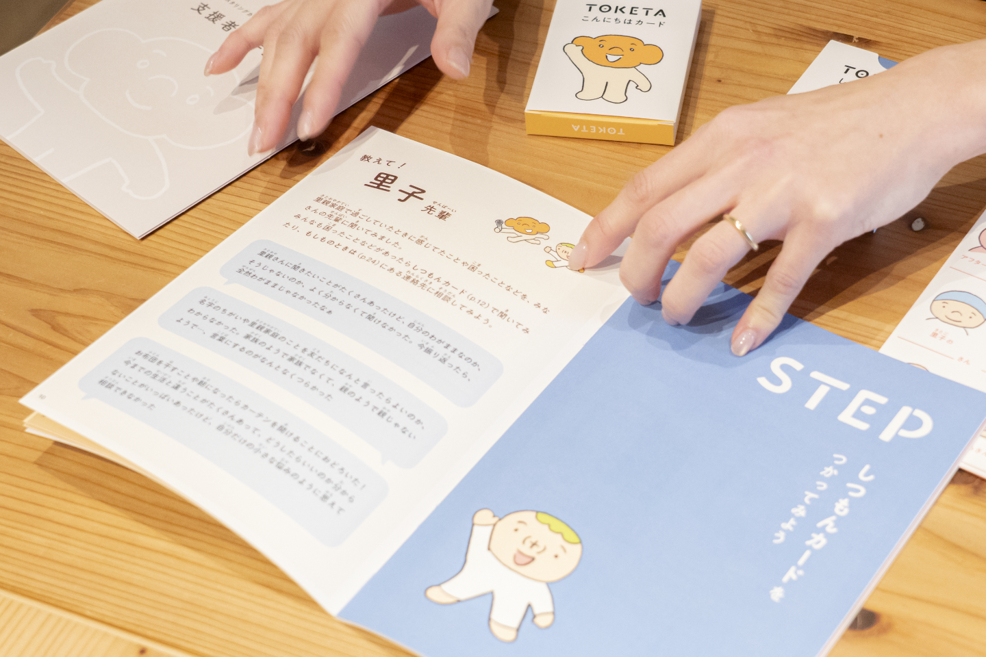 【写真】冊子とカードを扱いながら説明資料を開いている様子。「里子先輩」と書かれたページやイラストが見える