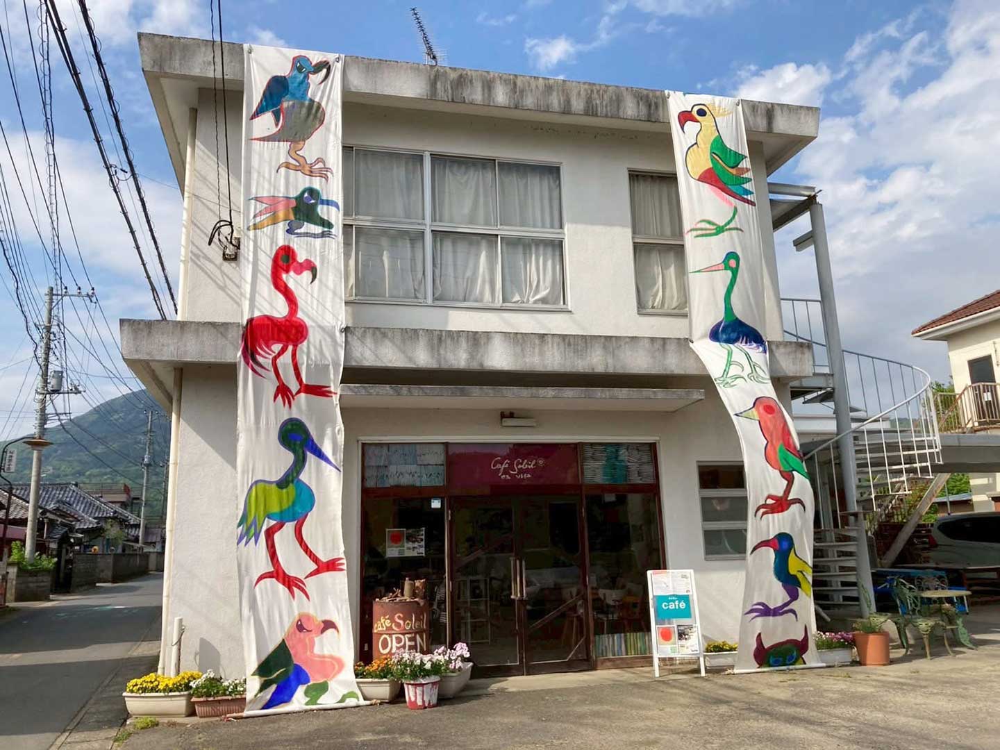 カラフルな鳥が描かれた幟が目を引く〈カフェソレイユ〉の外観写真。