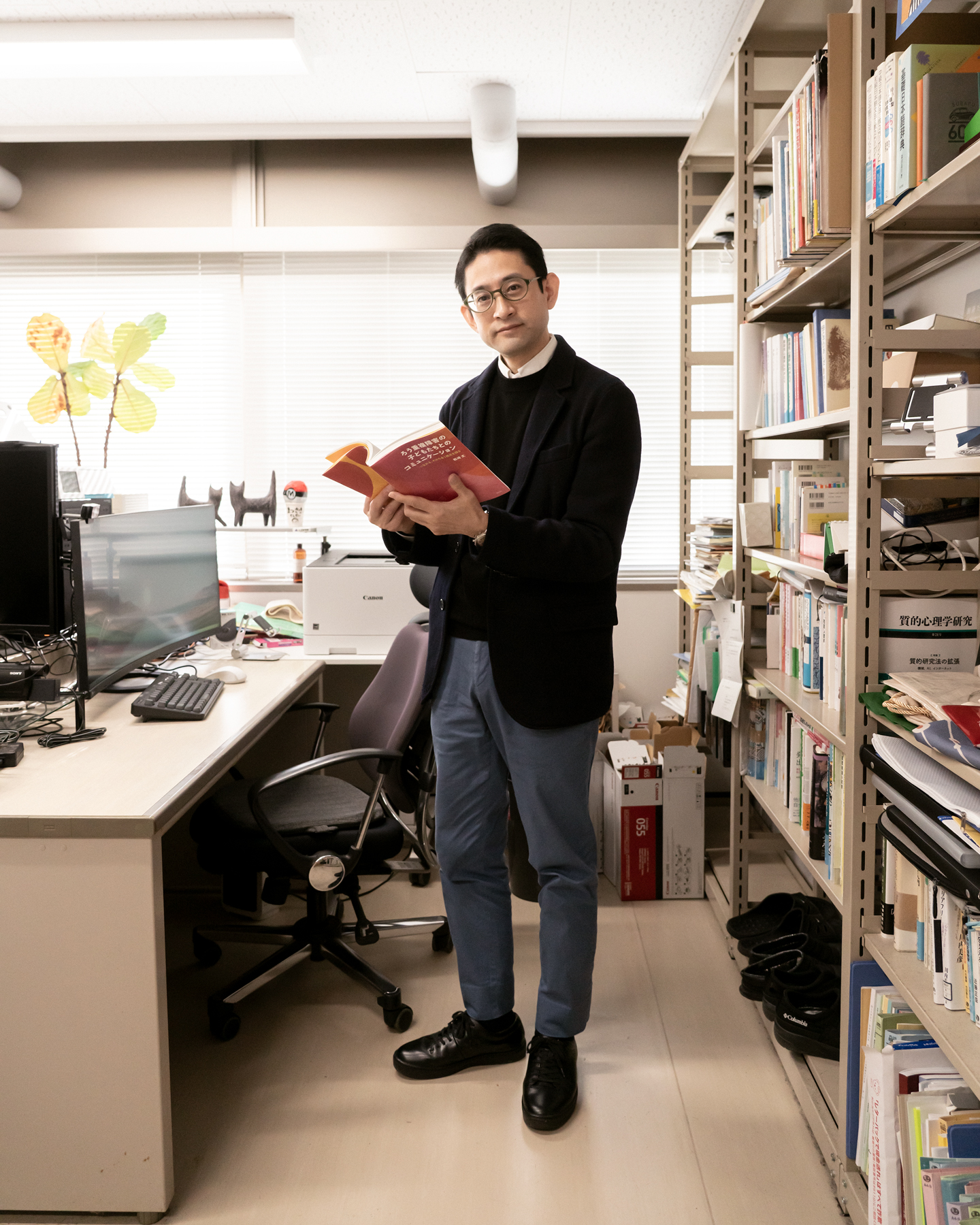 【写真】室内に立ち、赤い表紙の本を開いて正面を見ているまつざきじょうさん。向かって左手にはパソコンの置かれたデスクと椅子があり、右手はスチールラックの中に本や資料がならんでいる。松﨑さんはメガネ、黒いジャケットの下に黒いセーターと白いシャツ、青い色のパンツ、黒い革靴を身に着けている。