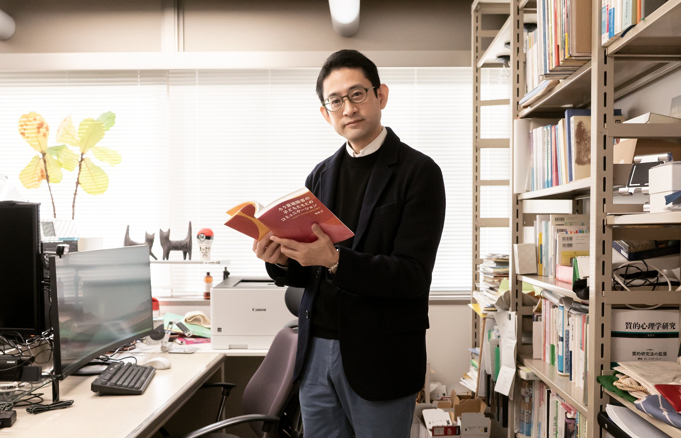 【写真】室内に立ち、赤い表紙の本を開いて正面を見ているまつざきじょうさん。向かって左手にはパソコンの置かれたデスクと椅子があり、右手はスチールラックの中に本や資料がならんでいる。松﨑さんはメガネ、黒いジャケットの下に黒いセーターと白いシャツ、青い色のパンツ、黒い革靴を身に着けている。