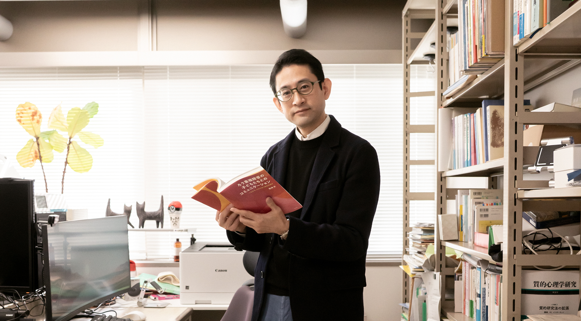 【写真】室内に立ち、赤い表紙の本を開いて正面を見ているまつざきじょうさん。向かって左手にはパソコンの置かれたデスクと椅子があり、右手はスチールラックの中に本や資料がならんでいる。松﨑さんはメガネ、黒いジャケットの下に黒いセーターと白いシャツ、青い色のパンツ、黒い革靴を身に着けている。