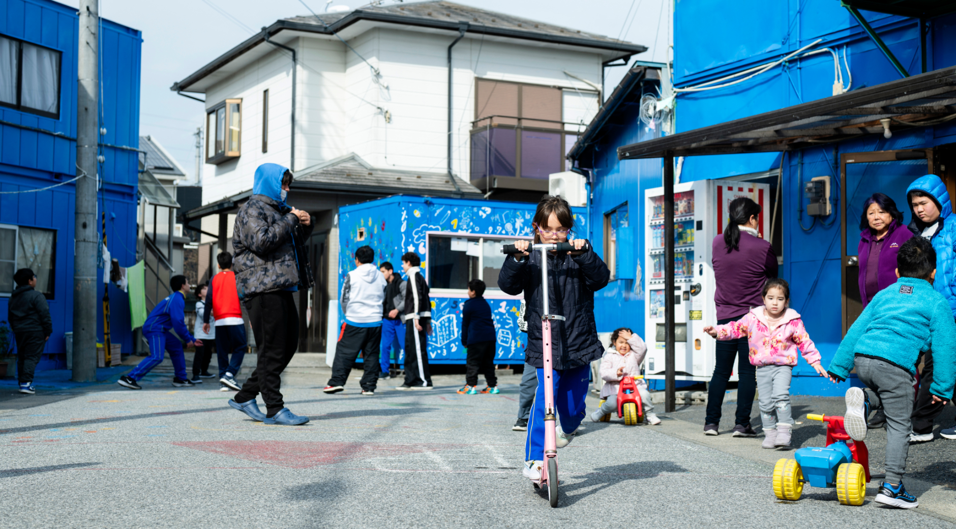 【写真】住宅街の青い建物の前で、たくさん子どもたち。中央では女の子がキックボードに乗り、他にもさまざまな年齢の子が思い思いにあそぶ