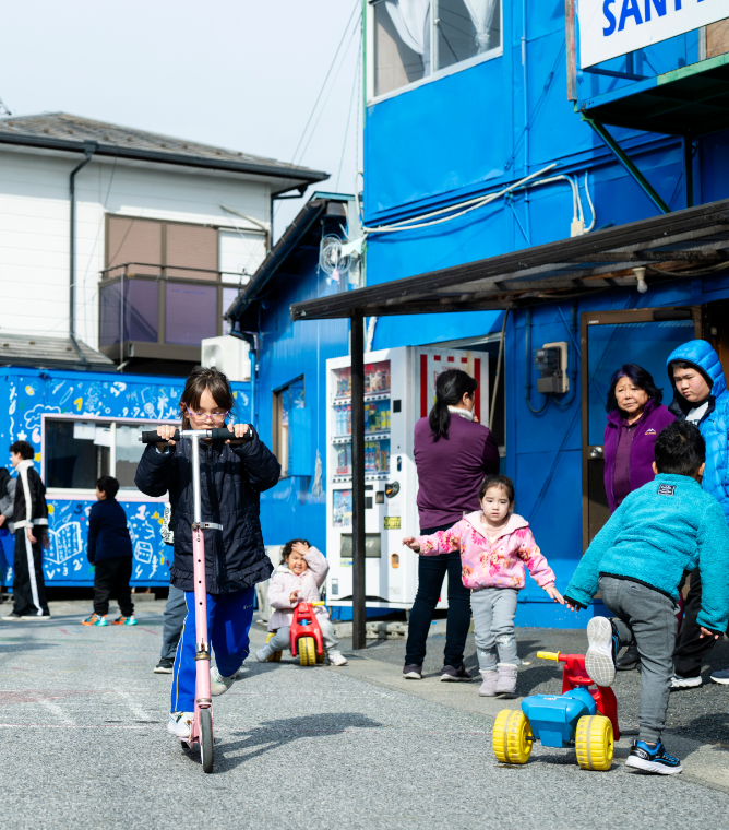 【写真】住宅街の青い建物の前で、たくさん子どもたち。中央では女の子がキックボードに乗り、他にもさまざまな年齢の子が思い思いにあそぶ