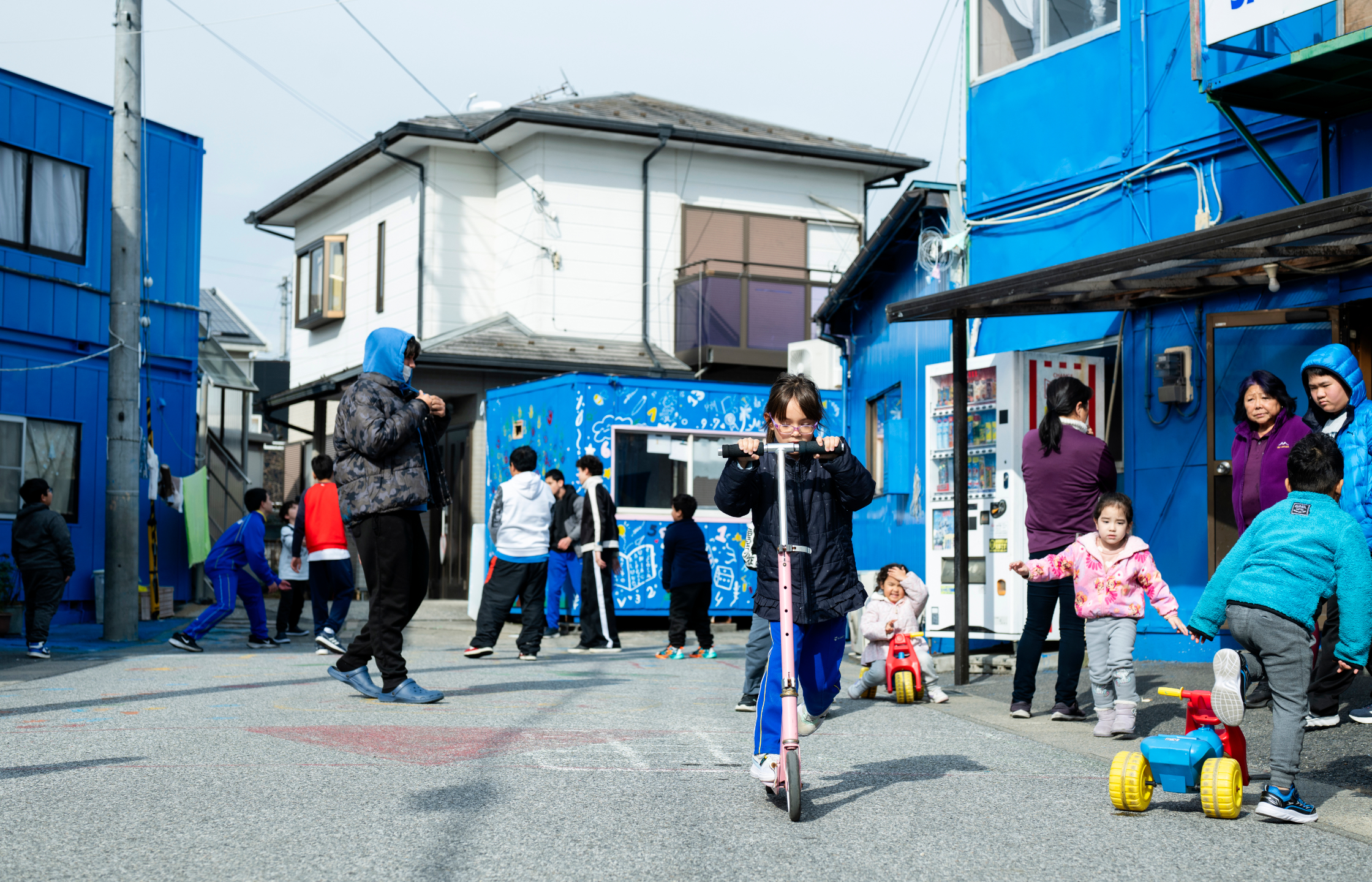 【写真】住宅街の青い建物の前で、たくさん子どもたち。中央では女の子がキックボードに乗り、他にもさまざまな年齢の子が思い思いにあそぶ