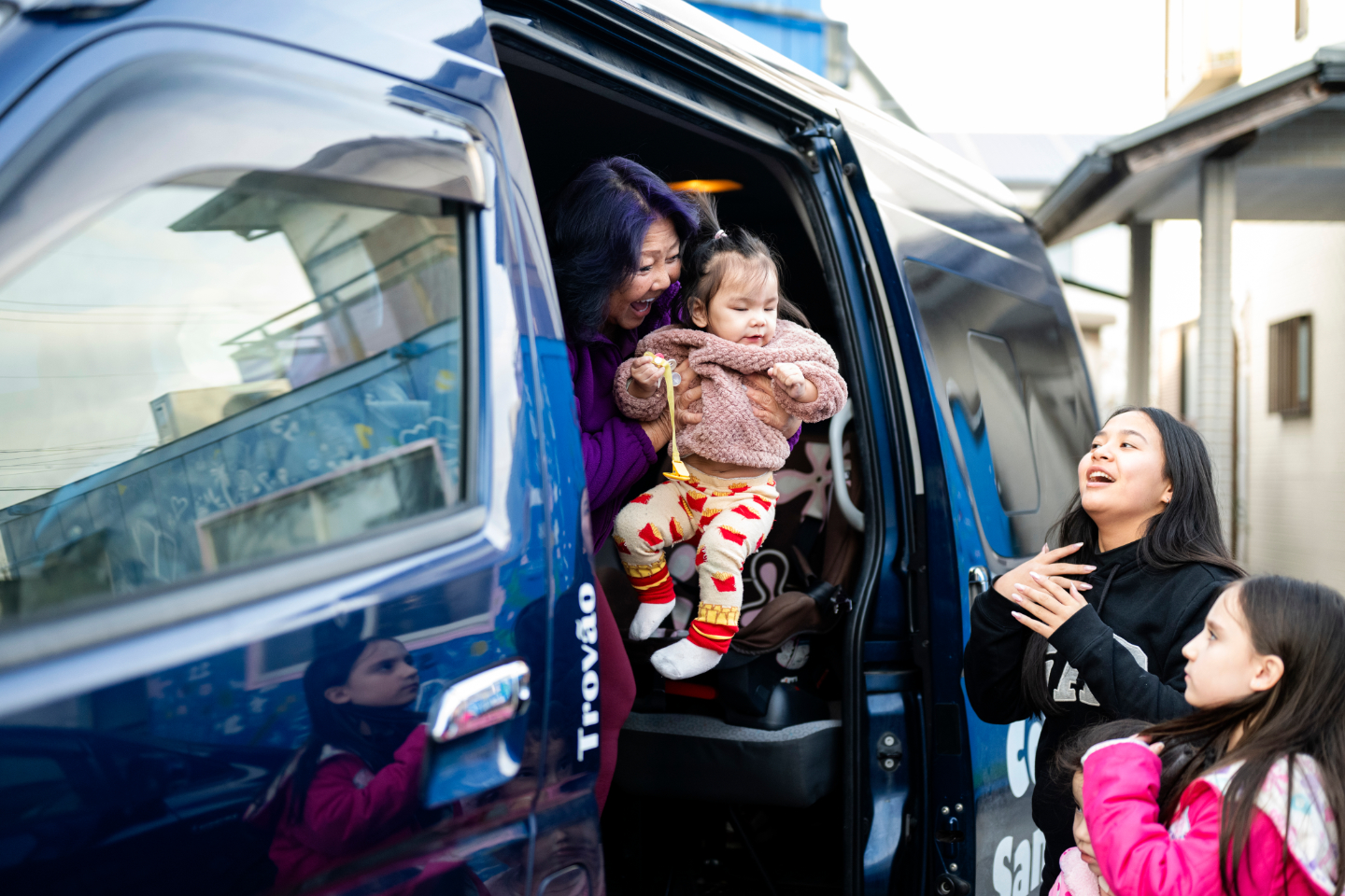 【写真】青い車のスライドドアから、ケンコ先生が幼い子どもを抱えて降ろしている