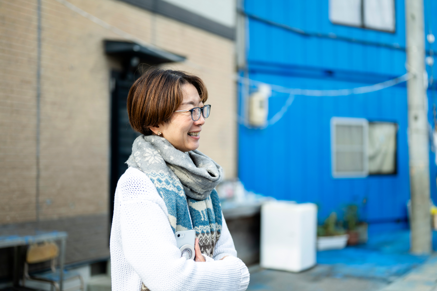 【写真】青い建物を背景に、マフラーを巻いた女性が笑顔で話を聞いている