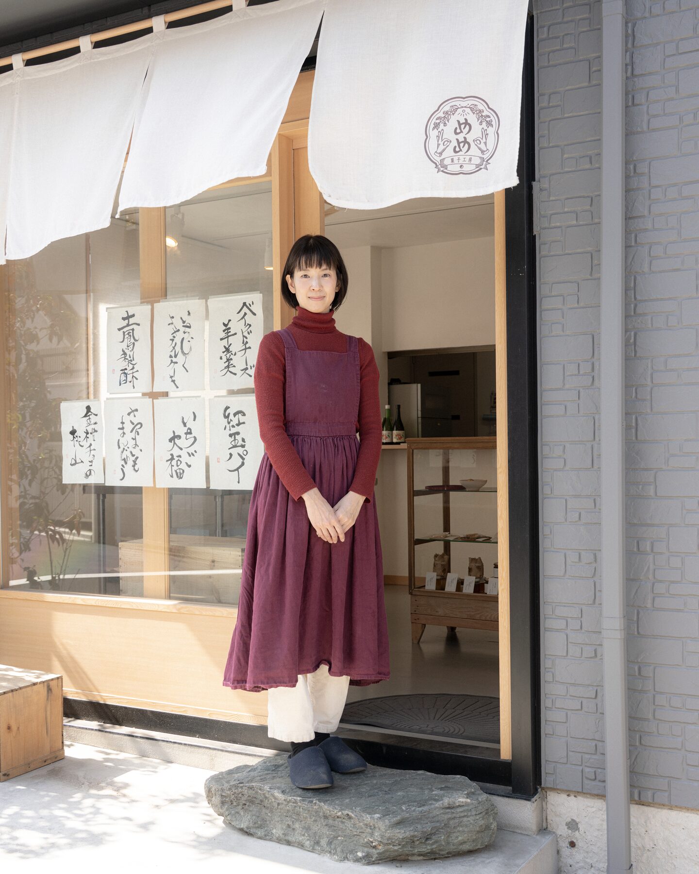【写真】えんじ色のワンピースの下に赤茶色の長袖、クリーム色のパンツを履いた伊藤ホサナさんが写真の何中に立ち、正面を向いている。店舗の前で、頭上にはお店のロゴが入った白いのれんがゆれている。ナホサさんの右肩の後ろには店舗のガラス窓があり、白い紙に黒い文字で販売しているお菓子の名前が書かれて張り出されている。