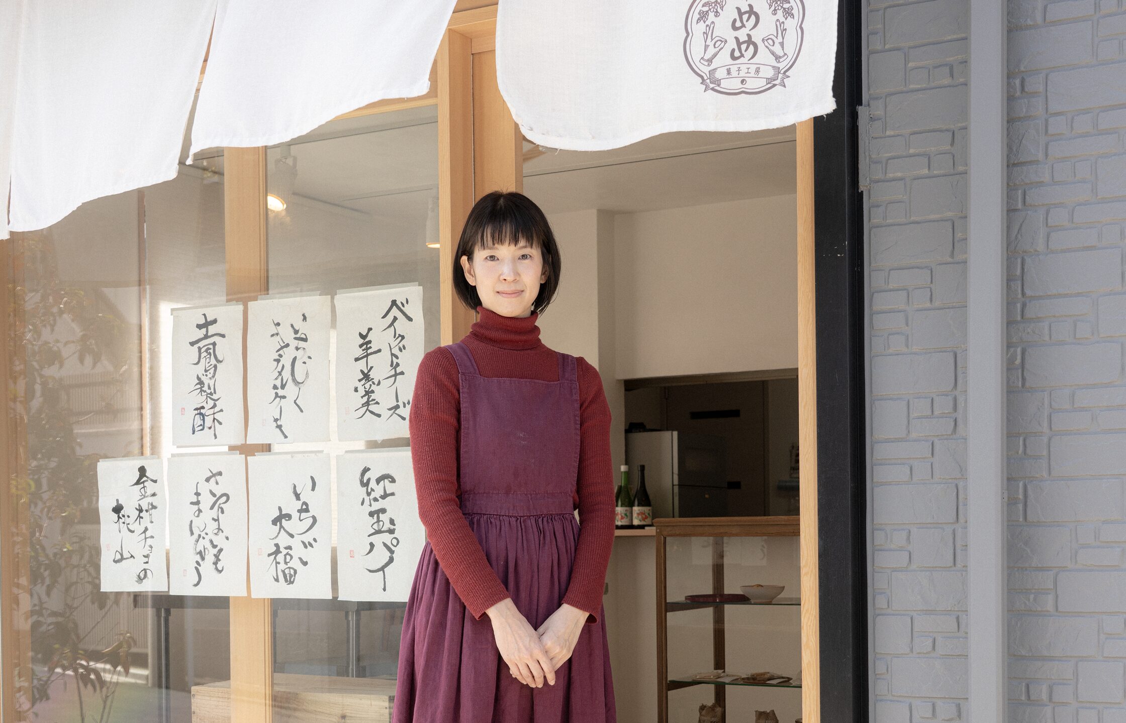 【写真】室内に立ち、赤い表紙の本を開いて正面を見ているまつざきじょうさん。向かって左手にはパソコンの置かれたデスクと椅子があり、右手はスチールラックの中に本や資料がならんでいる。松﨑さんはメガネ、黒いジャケットの下に黒いセーターと白いシャツ、青い色のパンツ、黒い革靴を身に着けている。