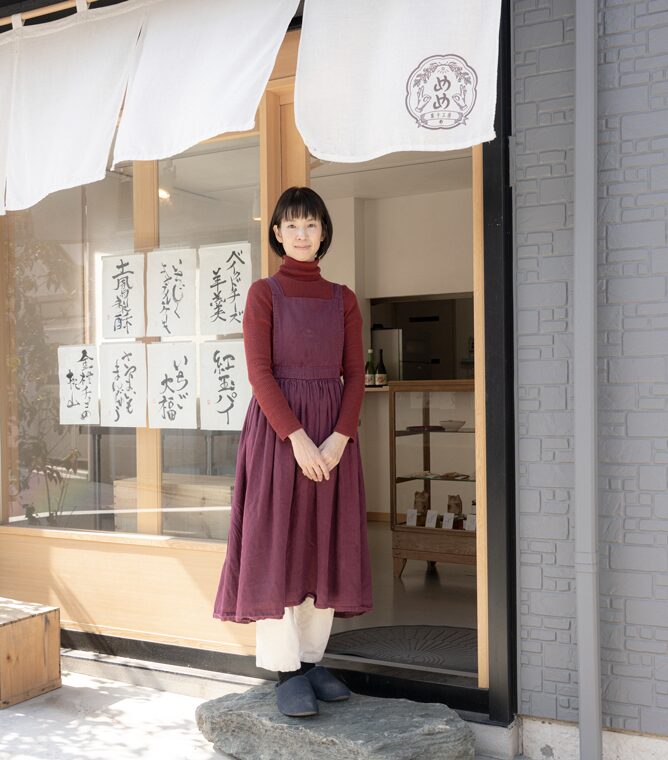 【写真】室内に立ち、赤い表紙の本を開いて正面を見ているまつざきじょうさん。向かって左手にはパソコンの置かれたデスクと椅子があり、右手はスチールラックの中に本や資料がならんでいる。松﨑さんはメガネ、黒いジャケットの下に黒いセーターと白いシャツ、青い色のパンツ、黒い革靴を身に着けている。