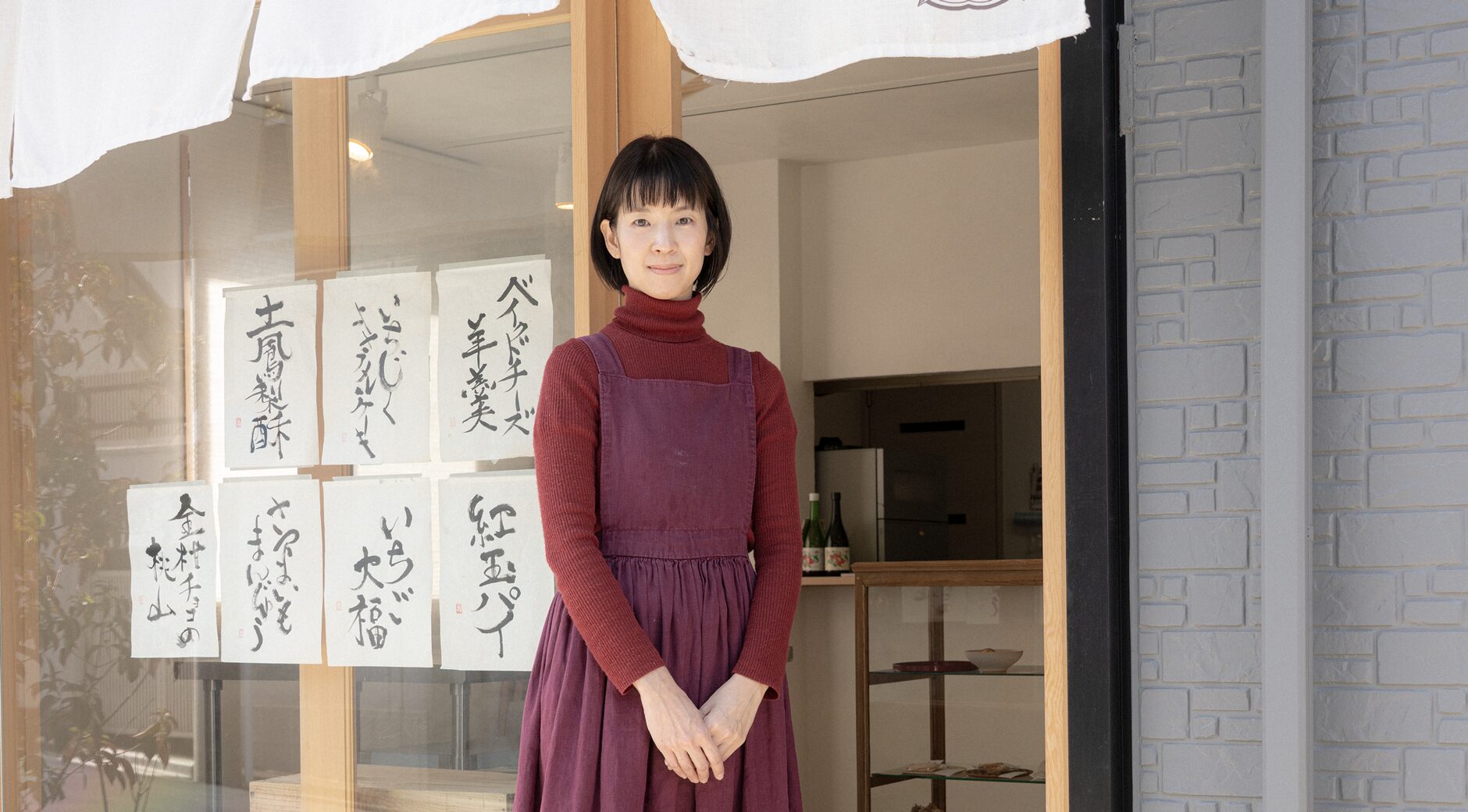 【写真】室内に立ち、赤い表紙の本を開いて正面を見ているまつざきじょうさん。向かって左手にはパソコンの置かれたデスクと椅子があり、右手はスチールラックの中に本や資料がならんでいる。松﨑さんはメガネ、黒いジャケットの下に黒いセーターと白いシャツ、青い色のパンツ、黒い革靴を身に着けている。