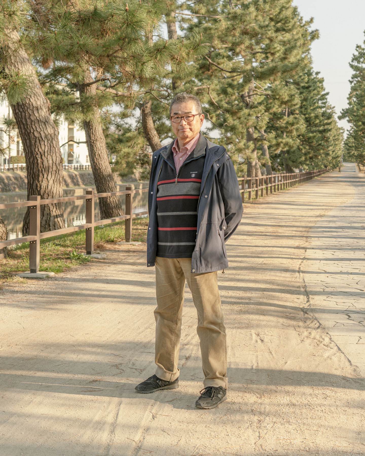 【写真】画面中央に宍戸さんが立っている。メガネをかけ、単発で、紺色のウィンドブレーカーの下にボーダーのセーターをきている。腕を後ろ手で組み、正面をみている。ししどさんのうしろにはまっすぐ歩道がのびてて、道沿いには松の木が並んでいる。