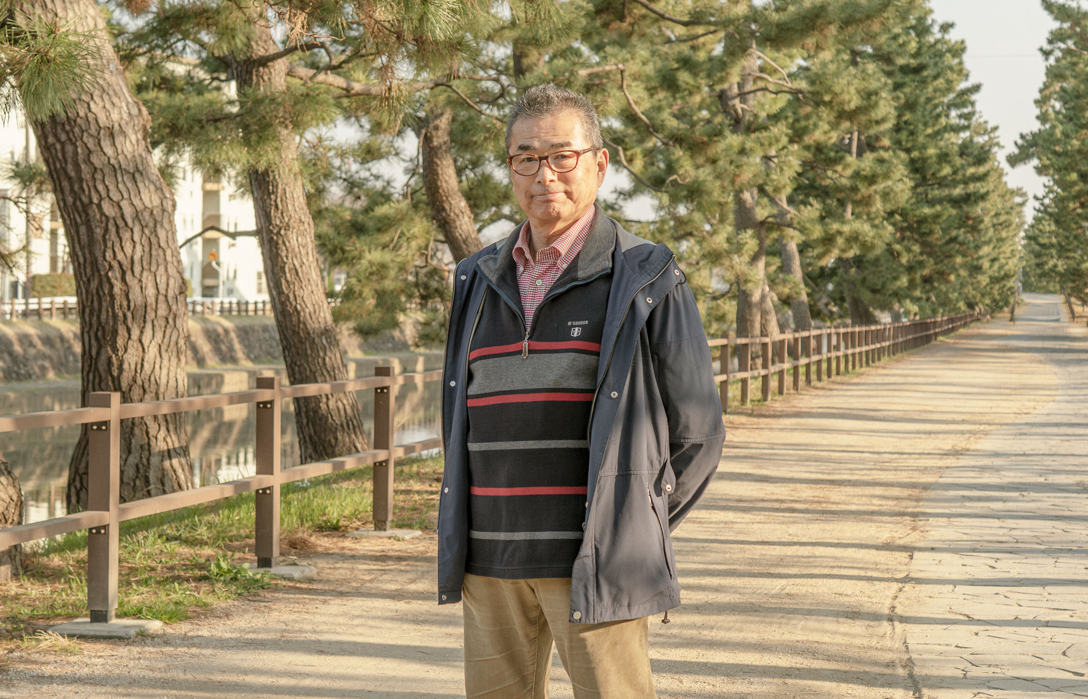 【写真】画面中央に宍戸さんが立っている。メガネをかけ、単発で、紺色のウィンドブレーカーの下にボーダーのセーターをきている。腕を後ろ手で組み、正面をみている。ししどさんのうしろにはまっすぐ歩道がのびてて、道沿いには松の木が並んでいる。