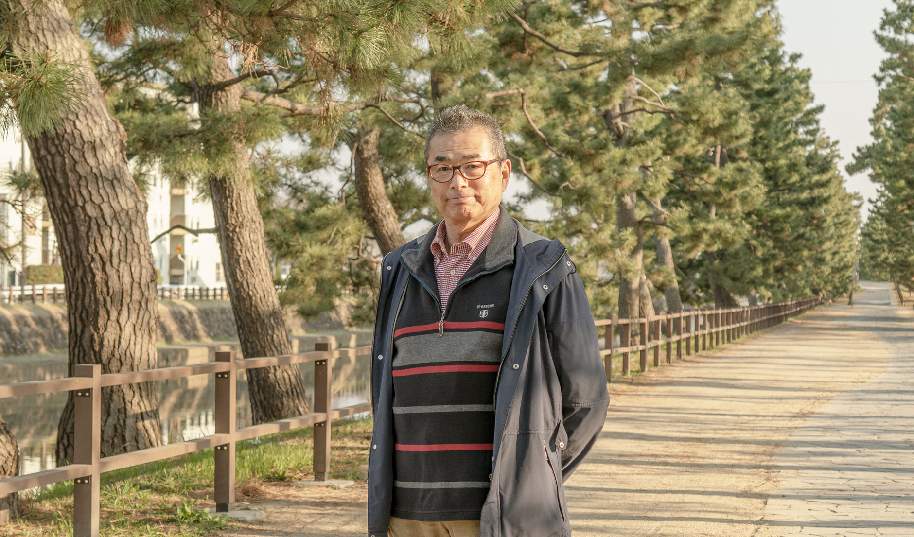 【写真】画面中央に宍戸さんが立っている。メガネをかけ、単発で、紺色のウィンドブレーカーの下にボーダーのセーターをきている。腕を後ろ手で組み、正面をみている。ししどさんのうしろにはまっすぐ歩道がのびてて、道沿いには松の木が並んでいる。