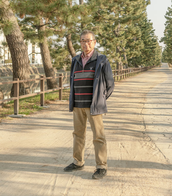 【写真】画面中央に宍戸さんが立っている。メガネをかけ、単発で、紺色のウィンドブレーカーの下にボーダーのセーターをきている。腕を後ろ手で組み、正面をみている。ししどさんのうしろにはまっすぐ歩道がのびてて、道沿いには松の木が並んでいる。