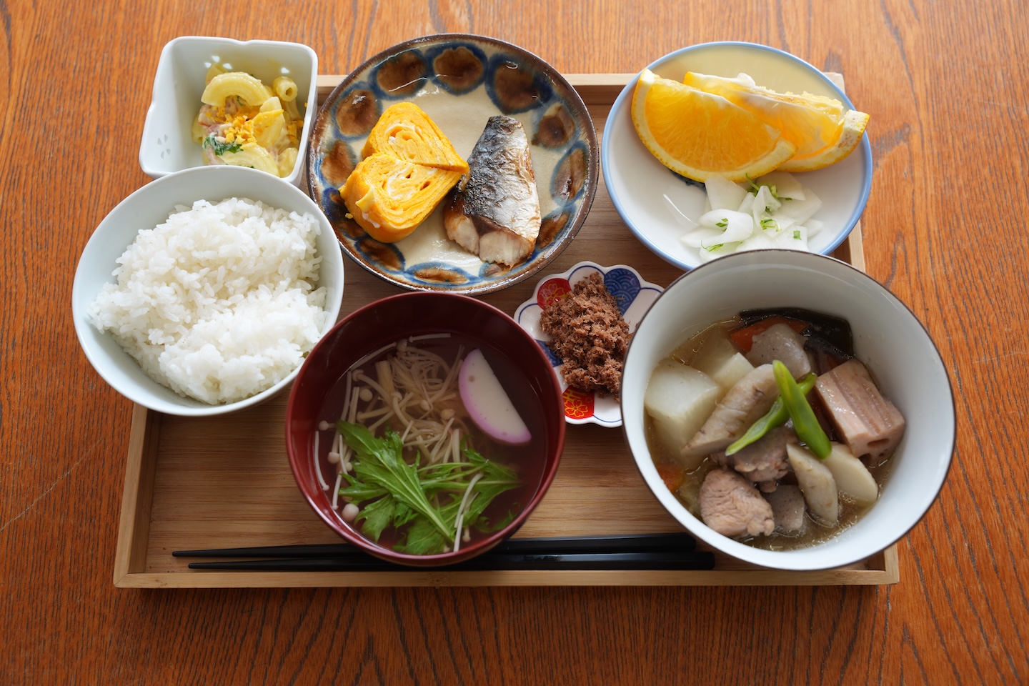 ごはんに汁物、煮物、焼き魚、卵焼き、果物などがのったプレートの写真