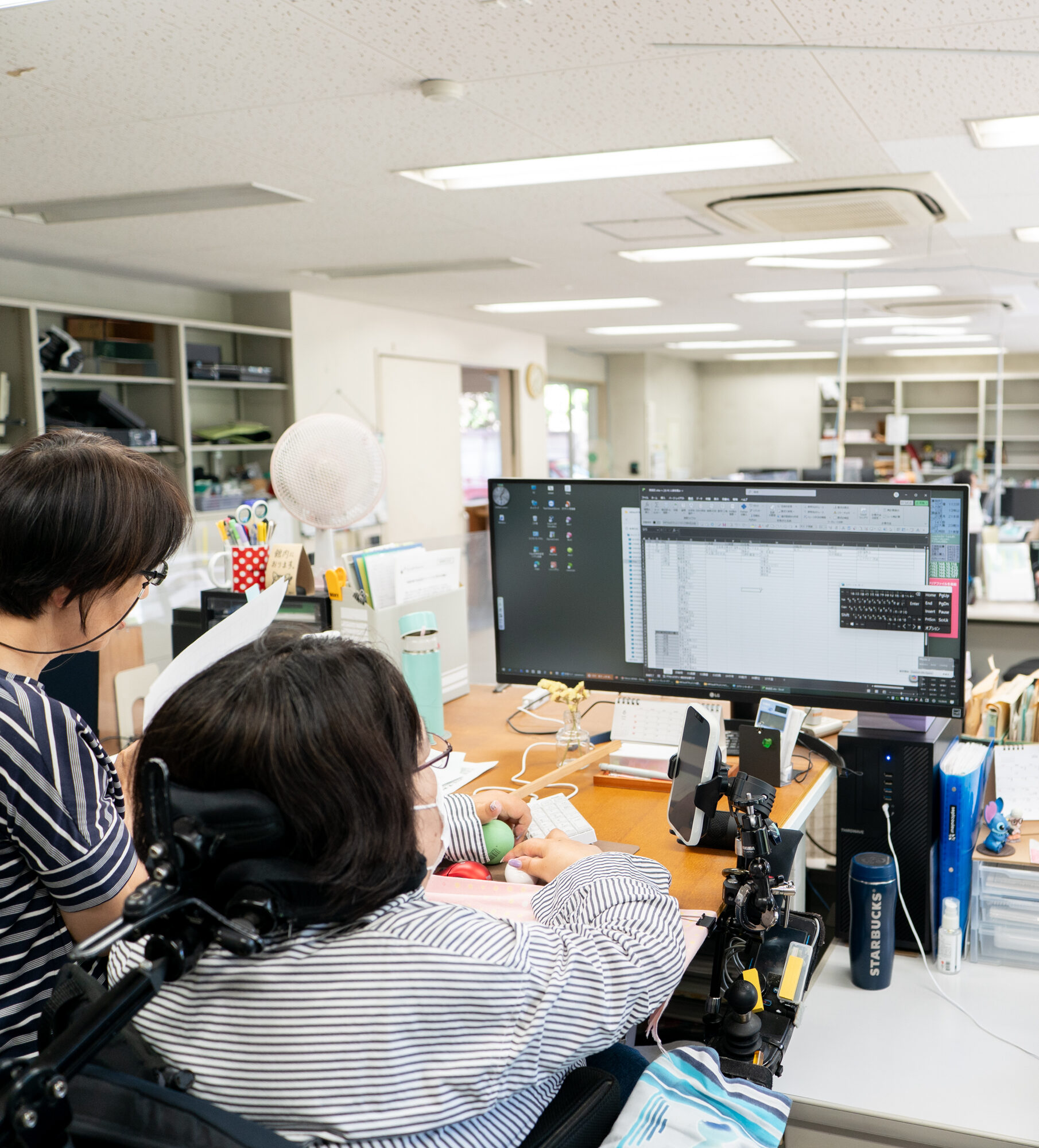 【写真】高めのデスクで作業をするひとと、そのそばで書類を眺めるひと